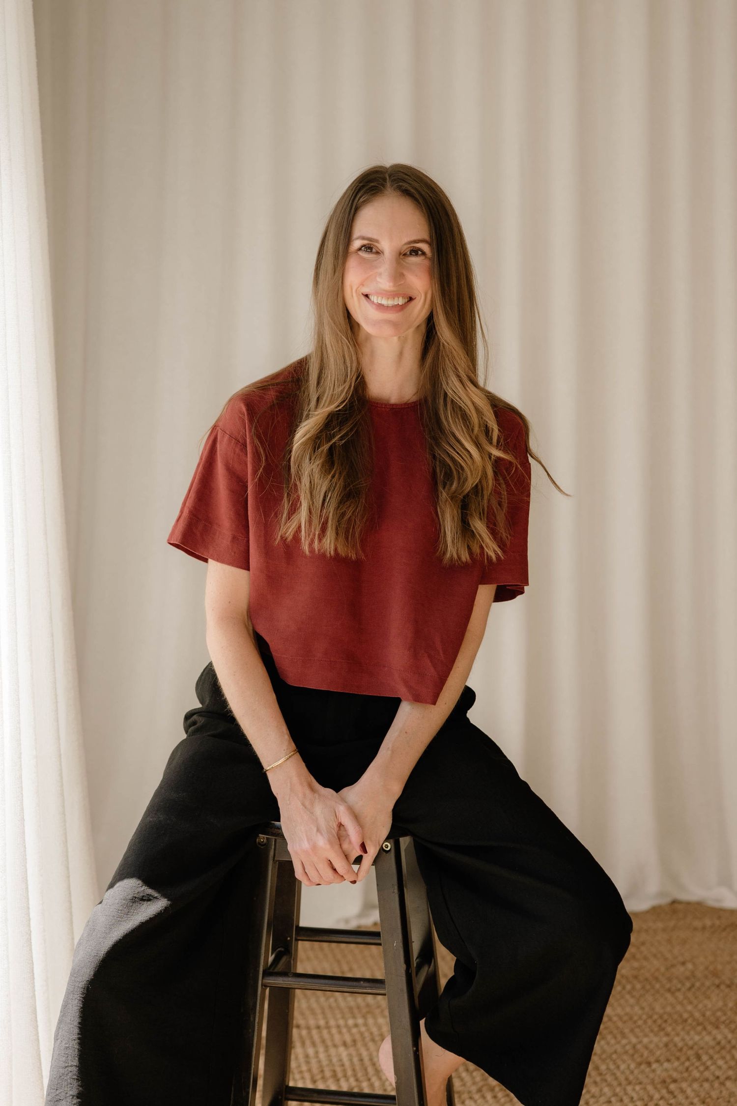 Confident woman in red crop top and black wide-leg pants sitting on stool smiling.