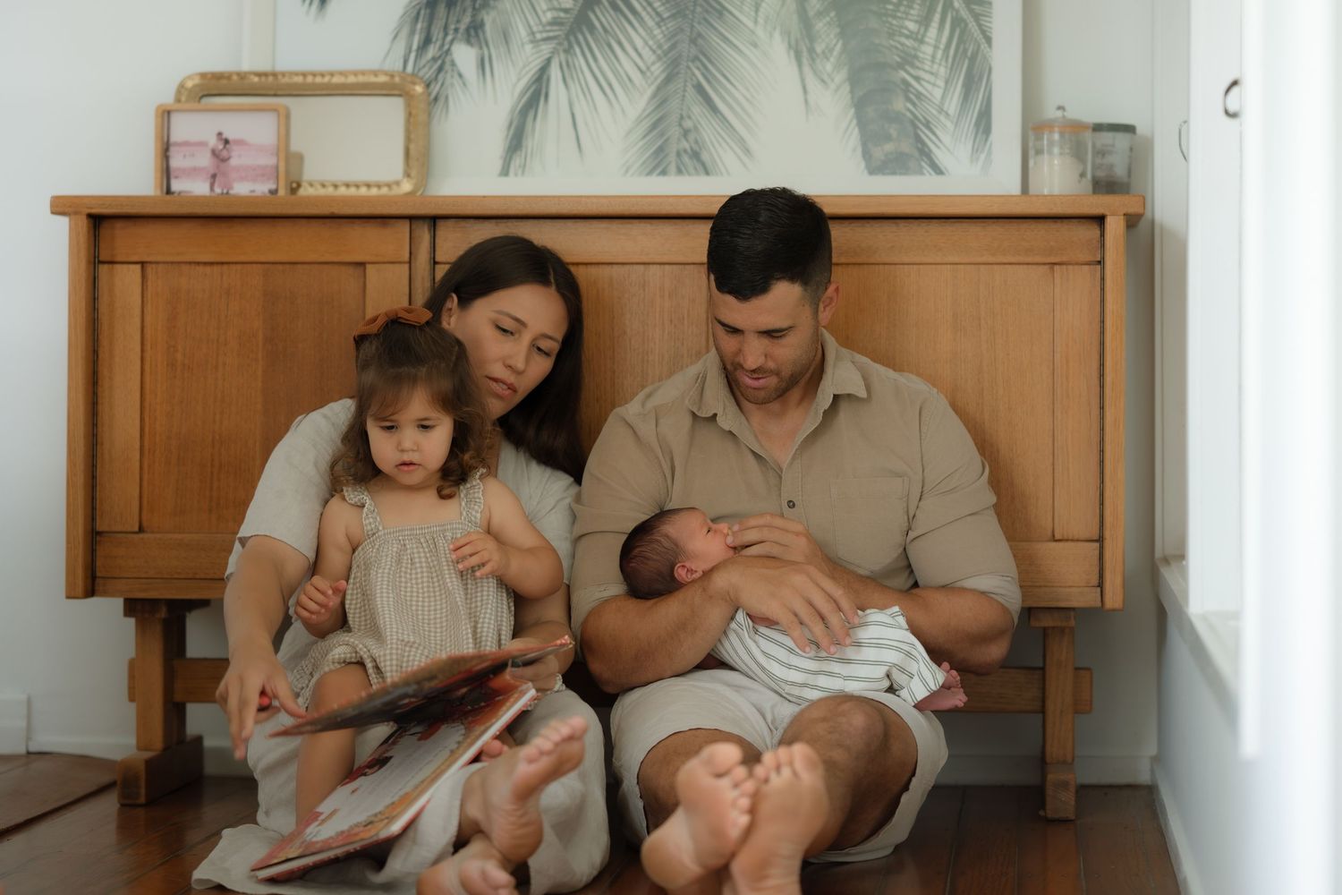 Family of four sitting by wooden dresser, parents reading to toddler while father holds newborn baby.