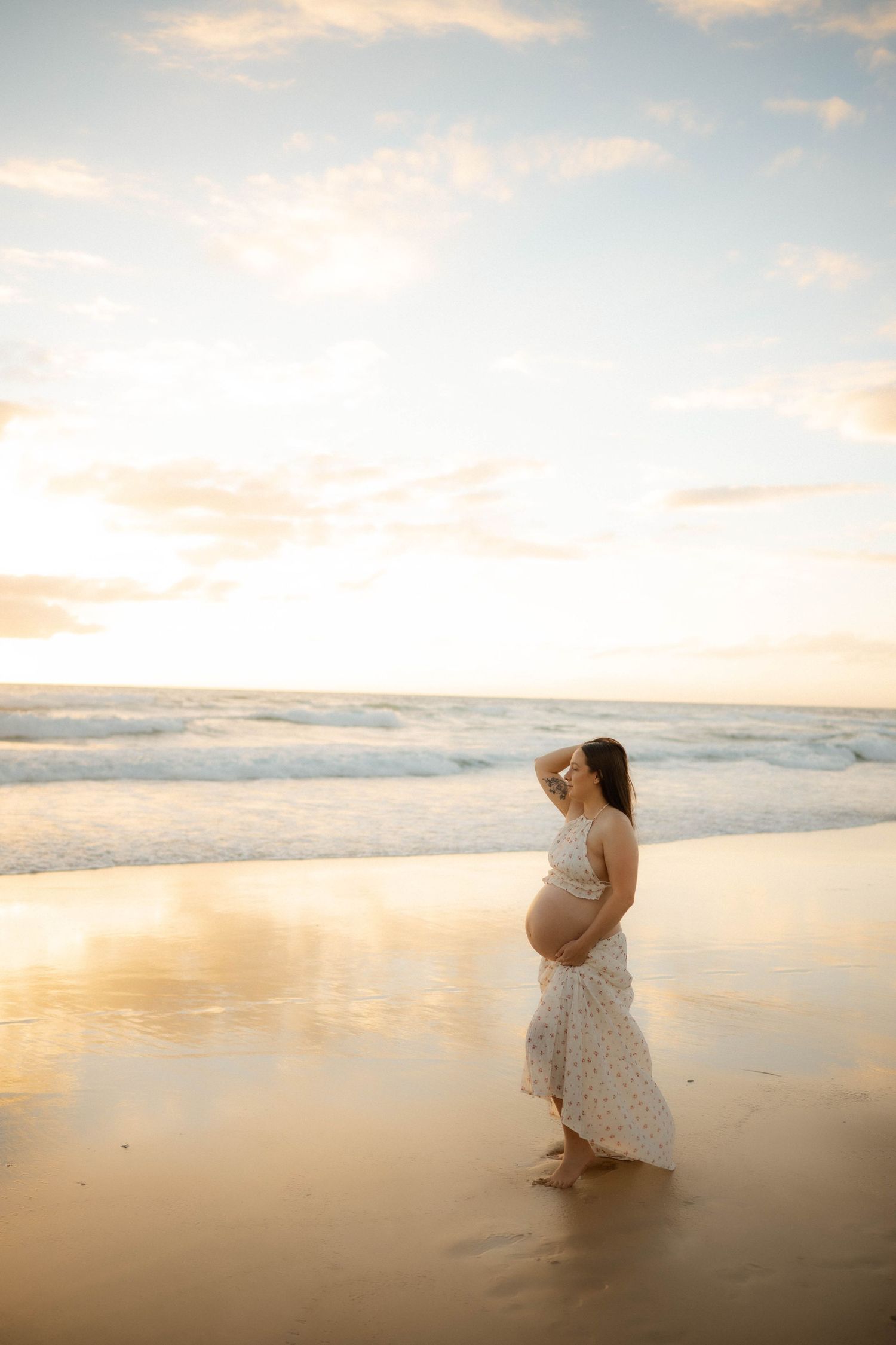 Barefoot pregnant woman in white floral dress gazing at ocean waves during a beautiful golden hour sunset.