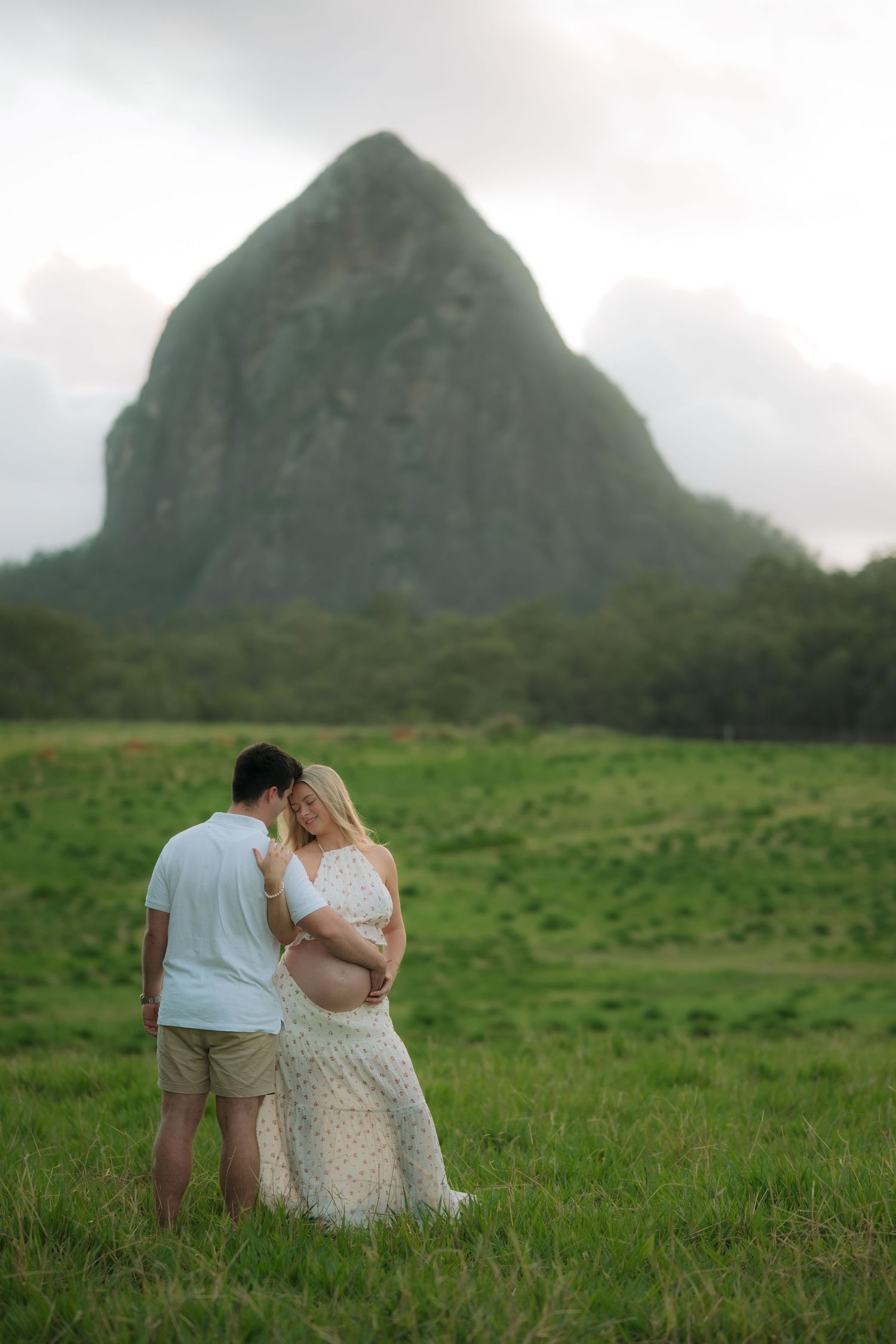 Pregnant couple embracing in green field with dramatic mountain peak backdrop at sunset.