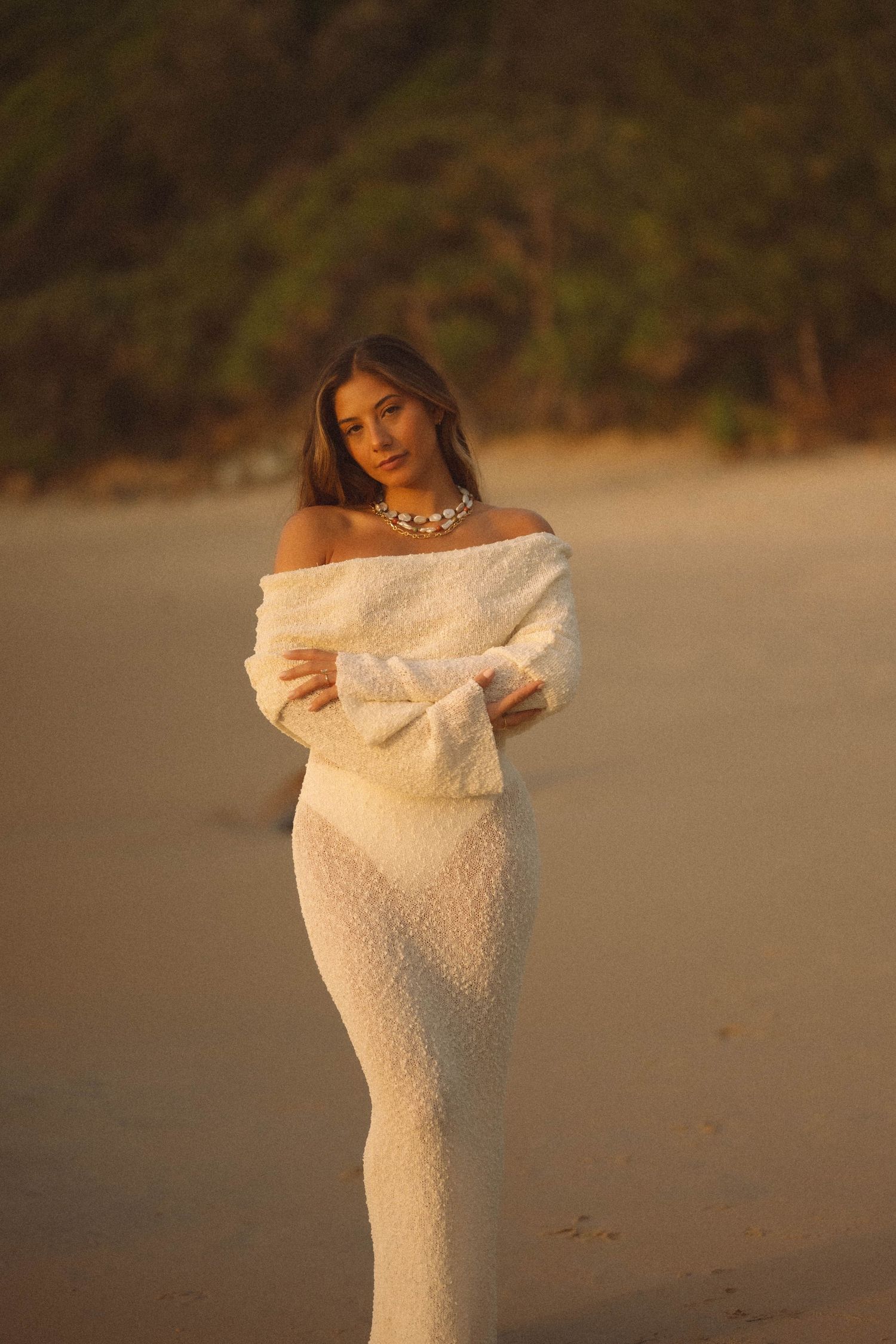 Woman in white off-shoulder textured dress poses on sandy beach at golden hour wearing a statement necklace.