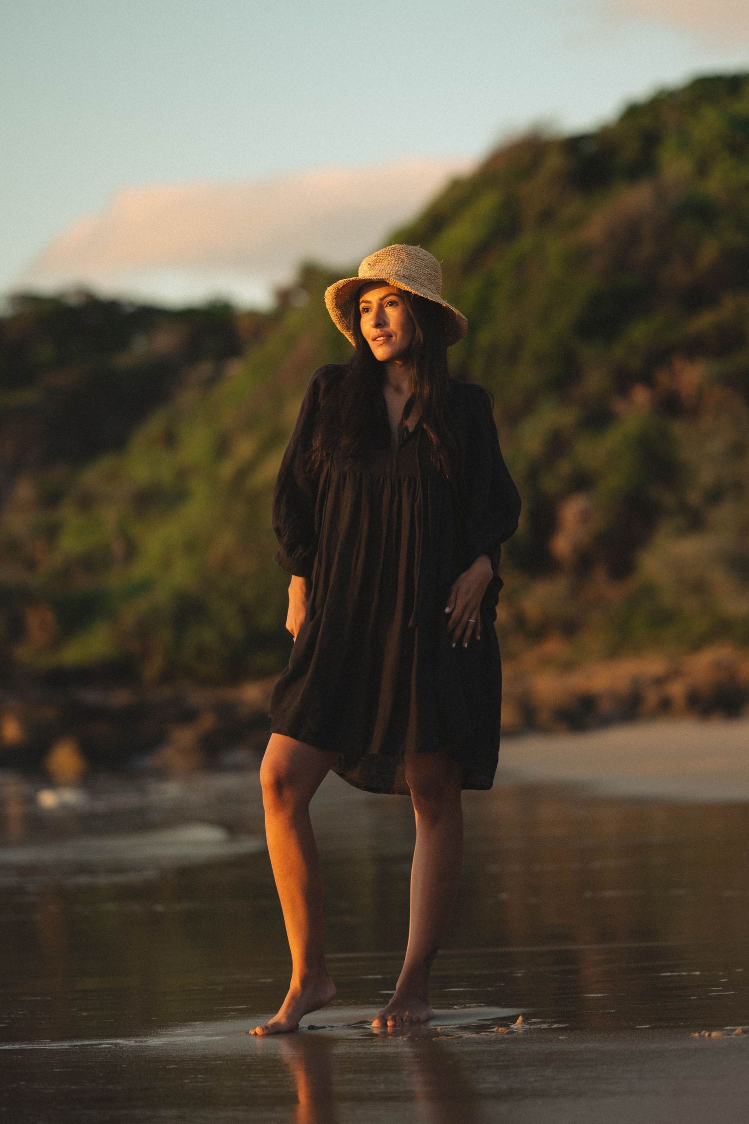 Woman in black dress and straw hat standing barefoot on a reflective beach at golden hour with green cliffs behind.