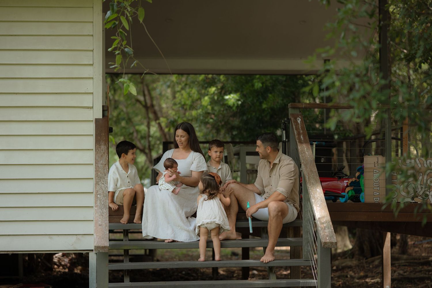 Family relaxing on rustic wooden porch steps with newborn baby, surrounded by natural green garden setting.
