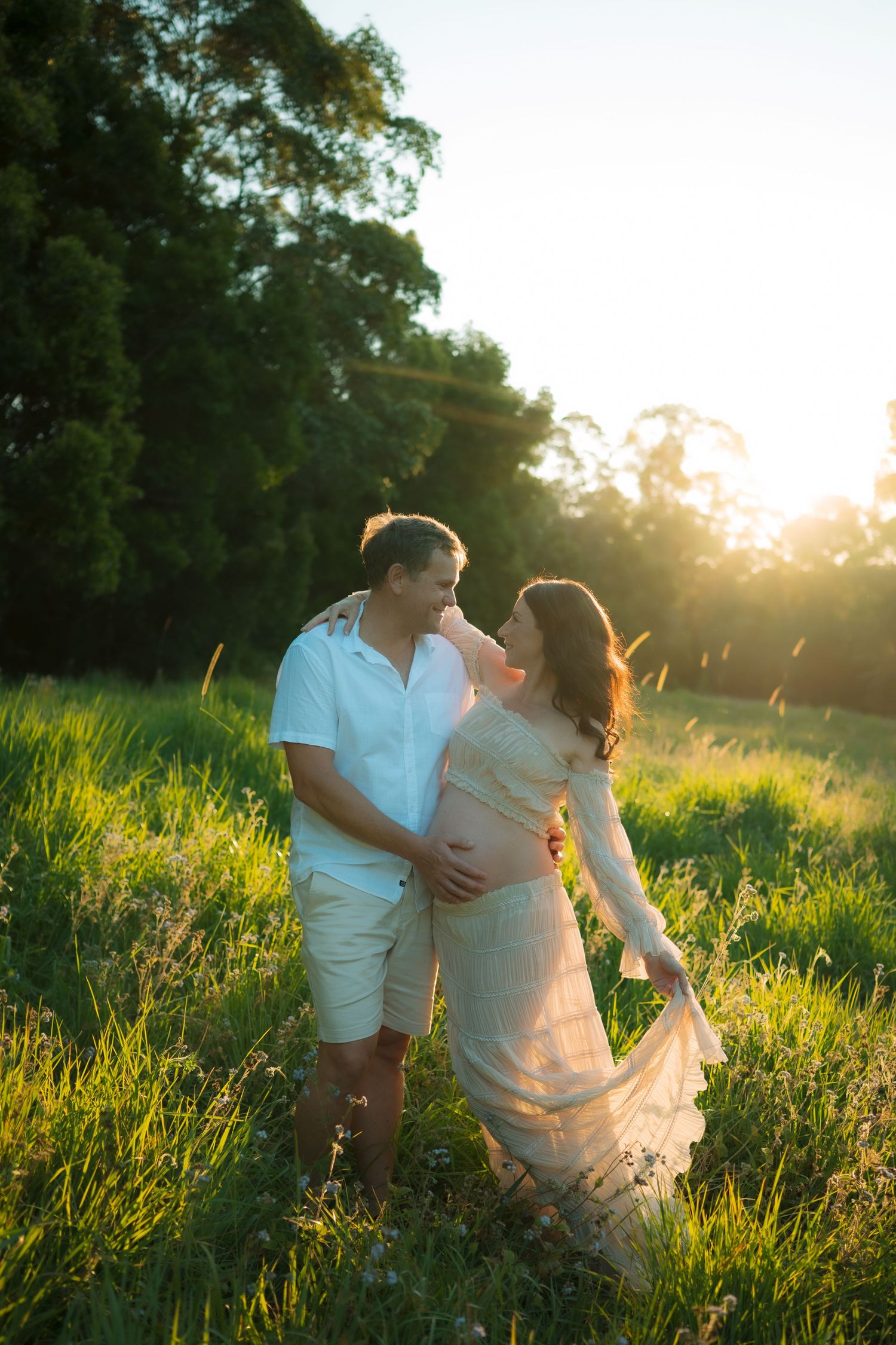 Happy expectant couple embraces in sunlit green meadow, man touching pregnant partners belly.