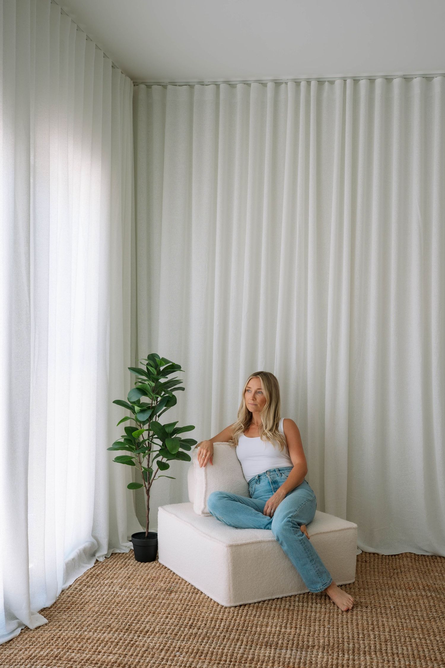 Relaxed woman in white tank and jeans sitting on ottoman beside fiddle leaf fig plant.