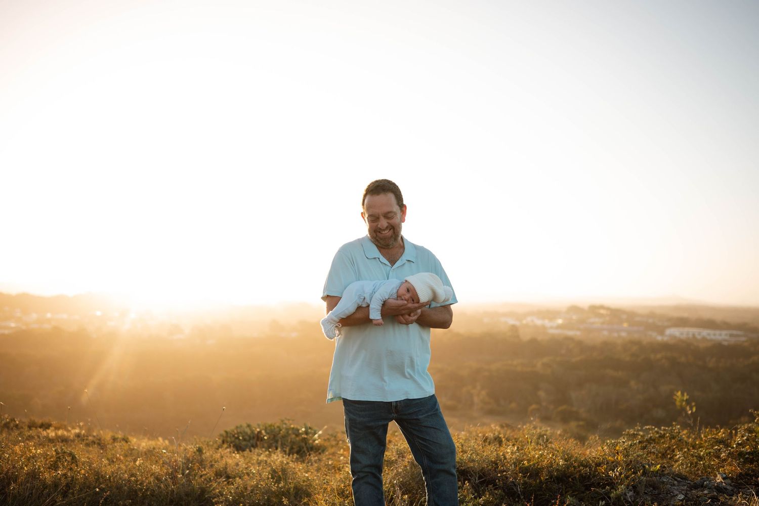 Man in light blue shirt smiling outdoors at golden sunset on hilltop.