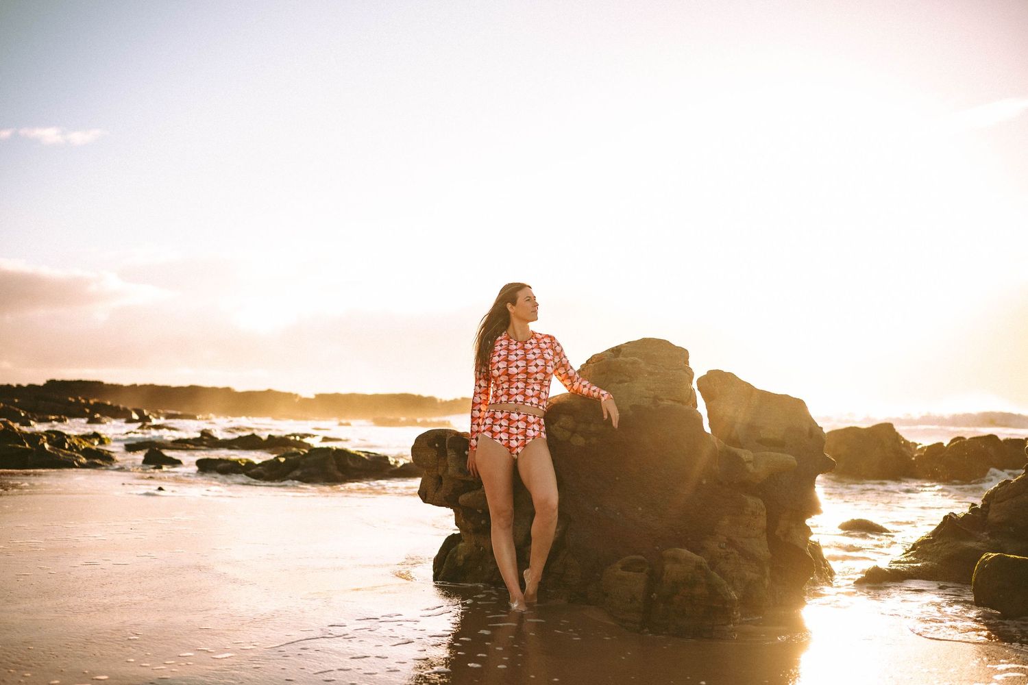 Woman in red floral shirt and white shorts leaning against coastal rocks at golden sunset beach.