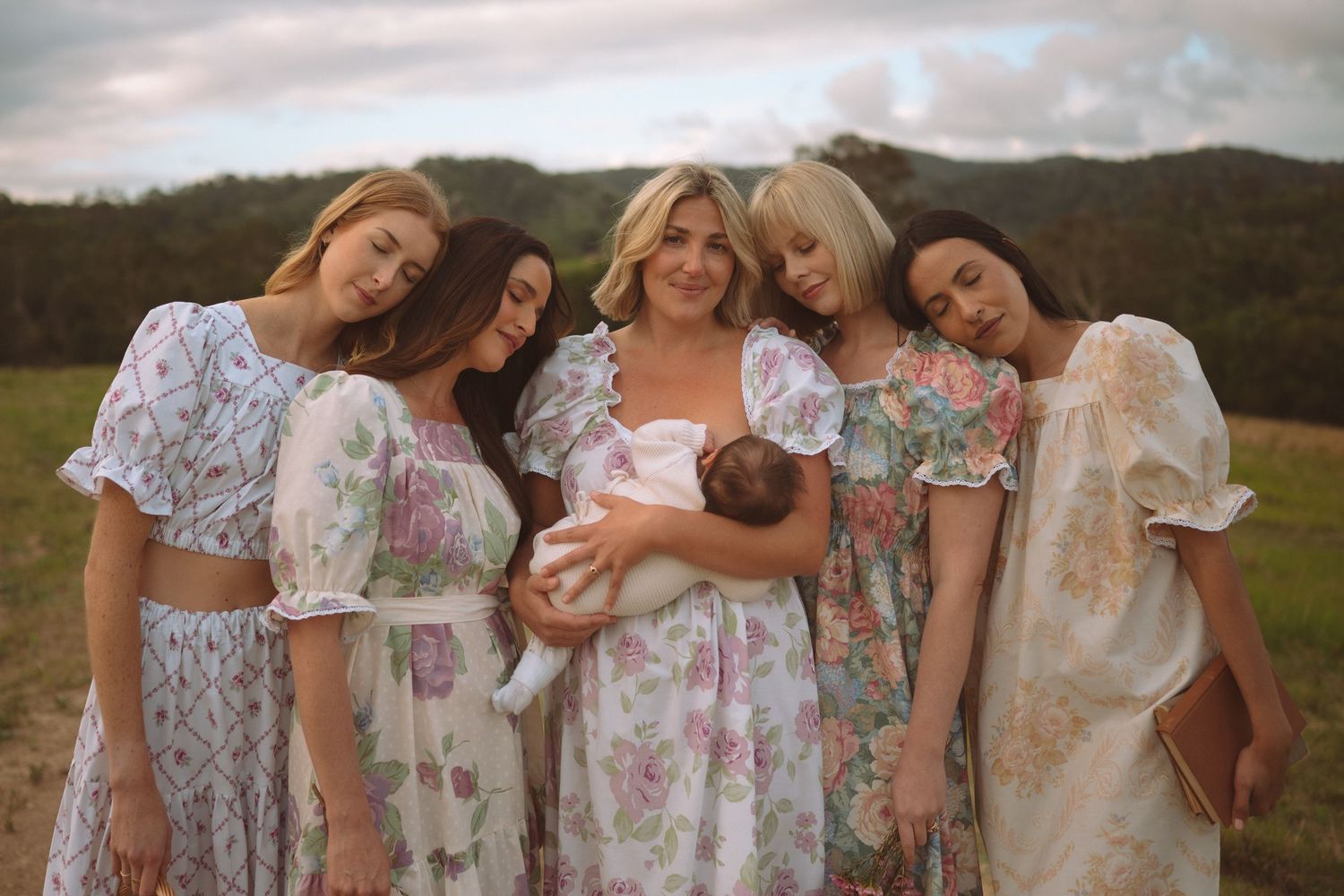 Five women in floral dresses gather outdoors, one holding a newborn baby, with green hills in the background.