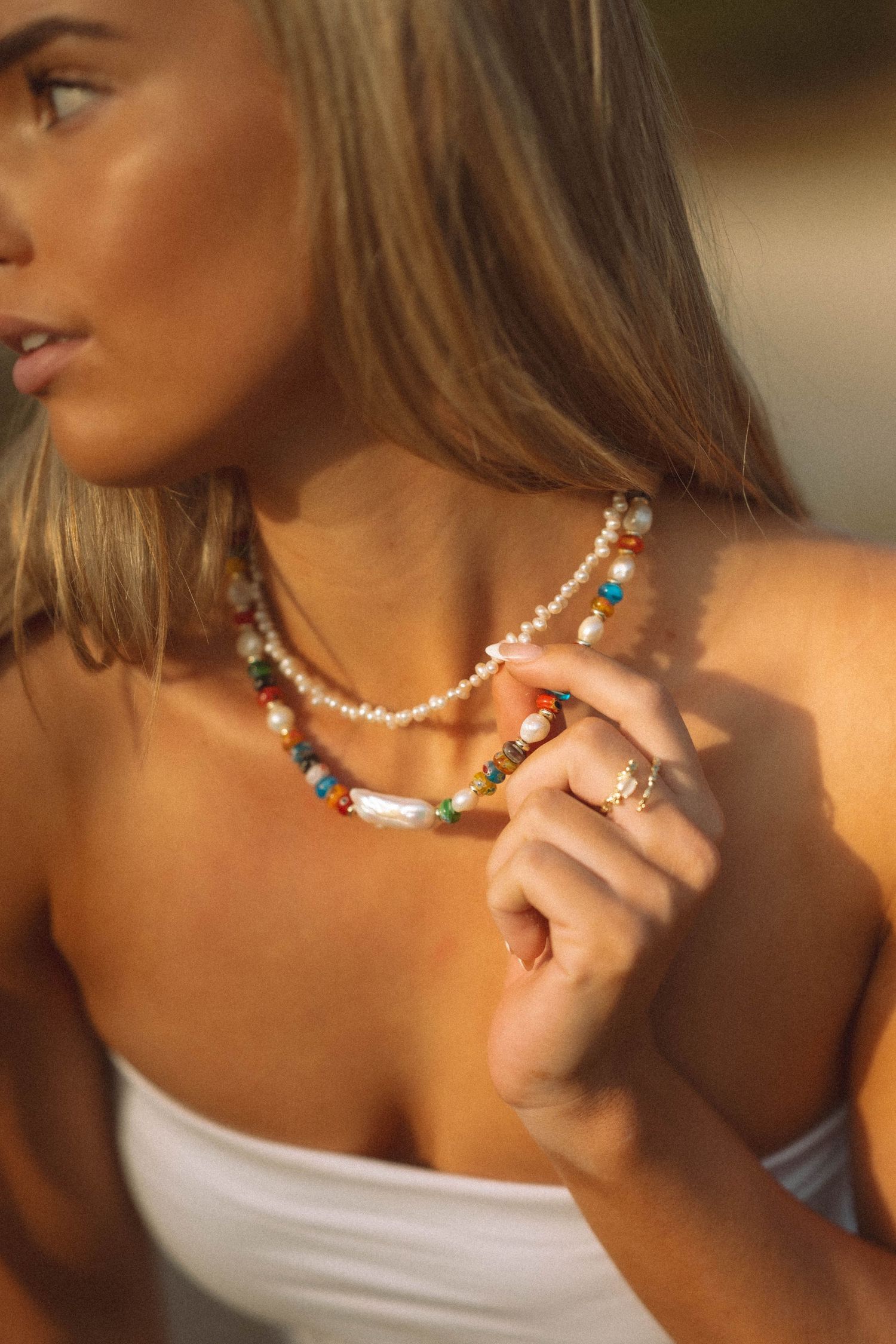 Woman wearing layered pearl and colorful beaded necklaces with a statement ring outdoors.