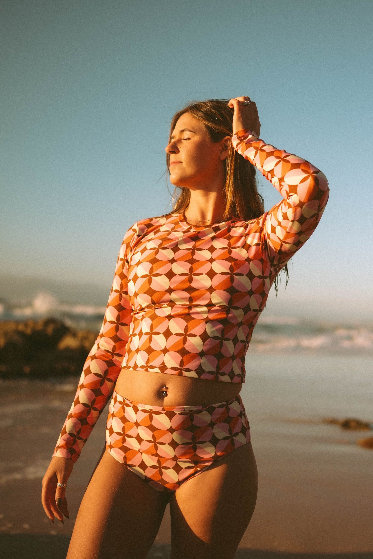 Woman in retro orange geometric print long-sleeve rashguard two-piece swimsuit on sunny beach.