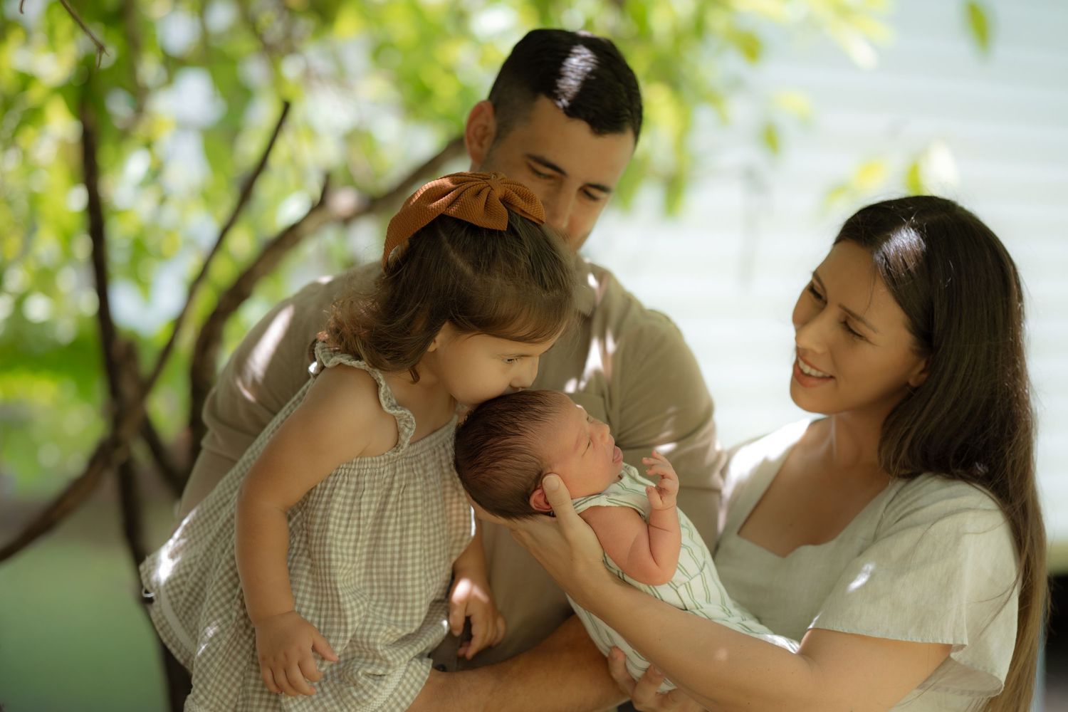 Parents and toddler daughter sharing tender moment with newborn baby outdoors under green leafy trees.
