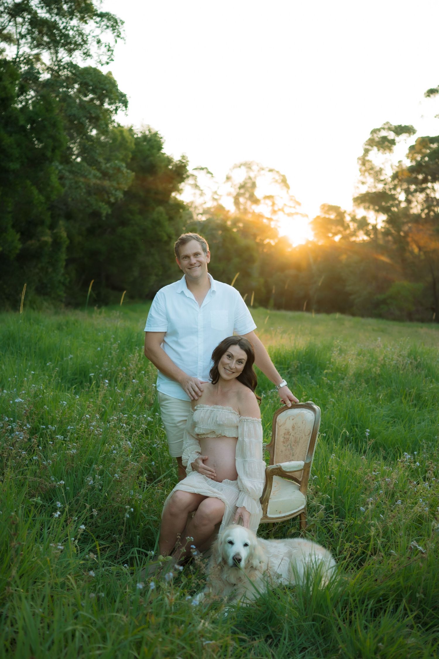 Smiling expectant couple poses with white dog and vintage chair in glowing sunset meadow.