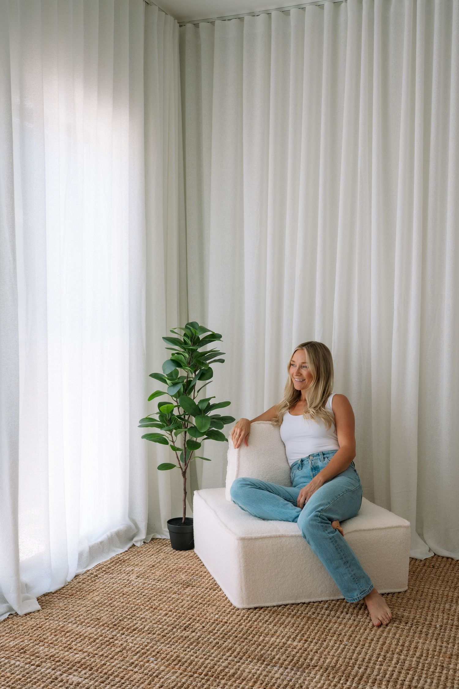 Blonde woman in white tank top and blue jeans sitting relaxed on ottoman beside fiddle leaf fig plant.