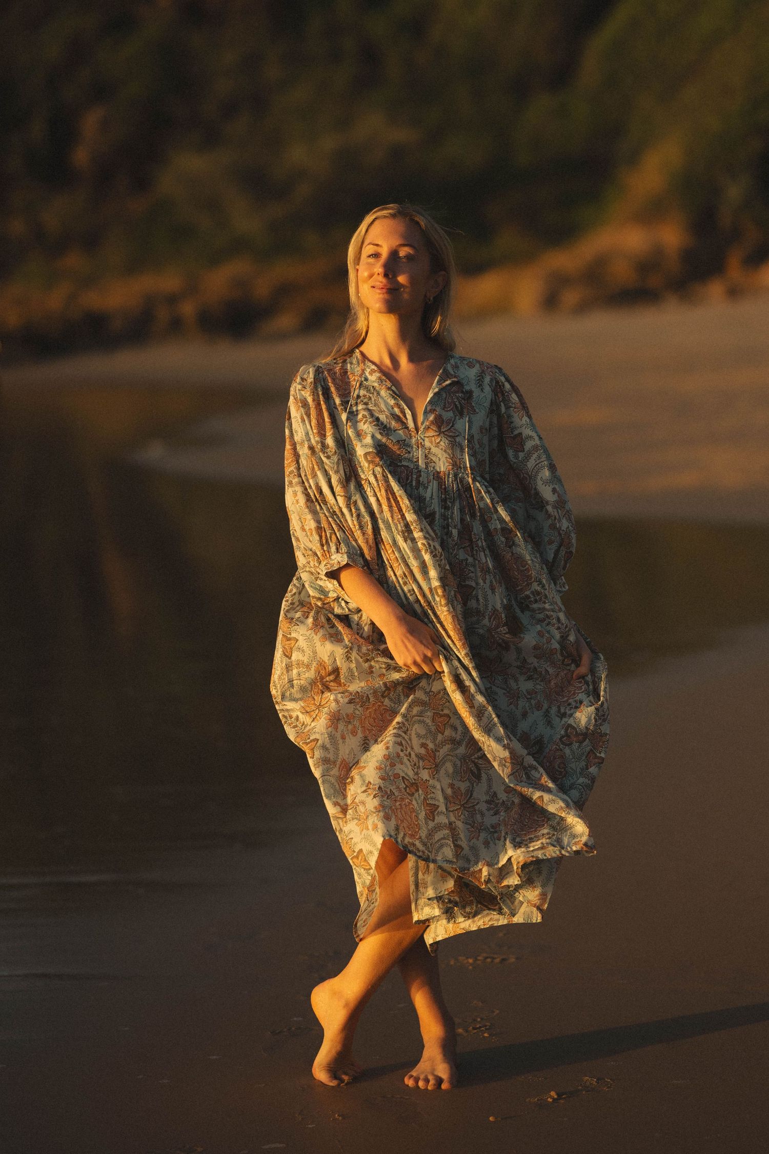 Woman in flowing floral kaftan walking barefoot on a sandy beach at golden sunset hour.