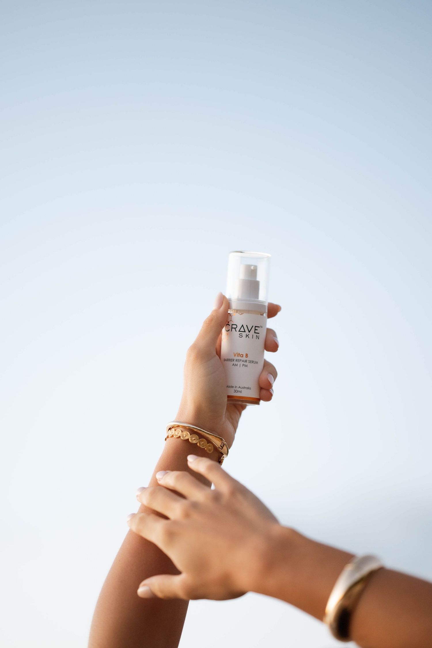 Hands holding a BRAVE skin care serum bottle against a clear blue sky, wearing gold bracelets.