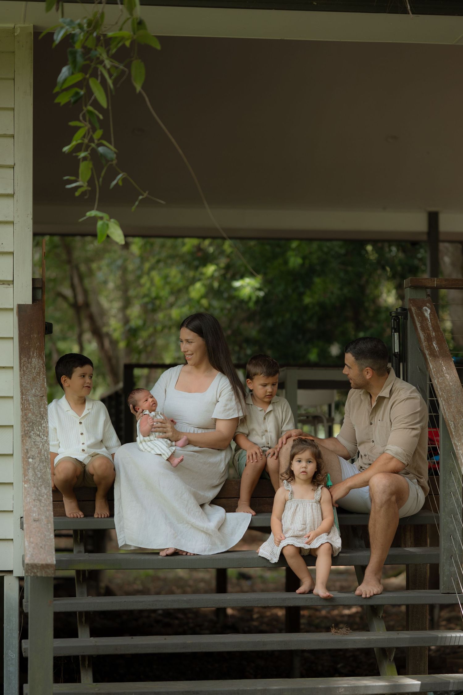 Family of six sitting on wooden porch steps surrounded by lush greenery, mother in white dress holding newborn.