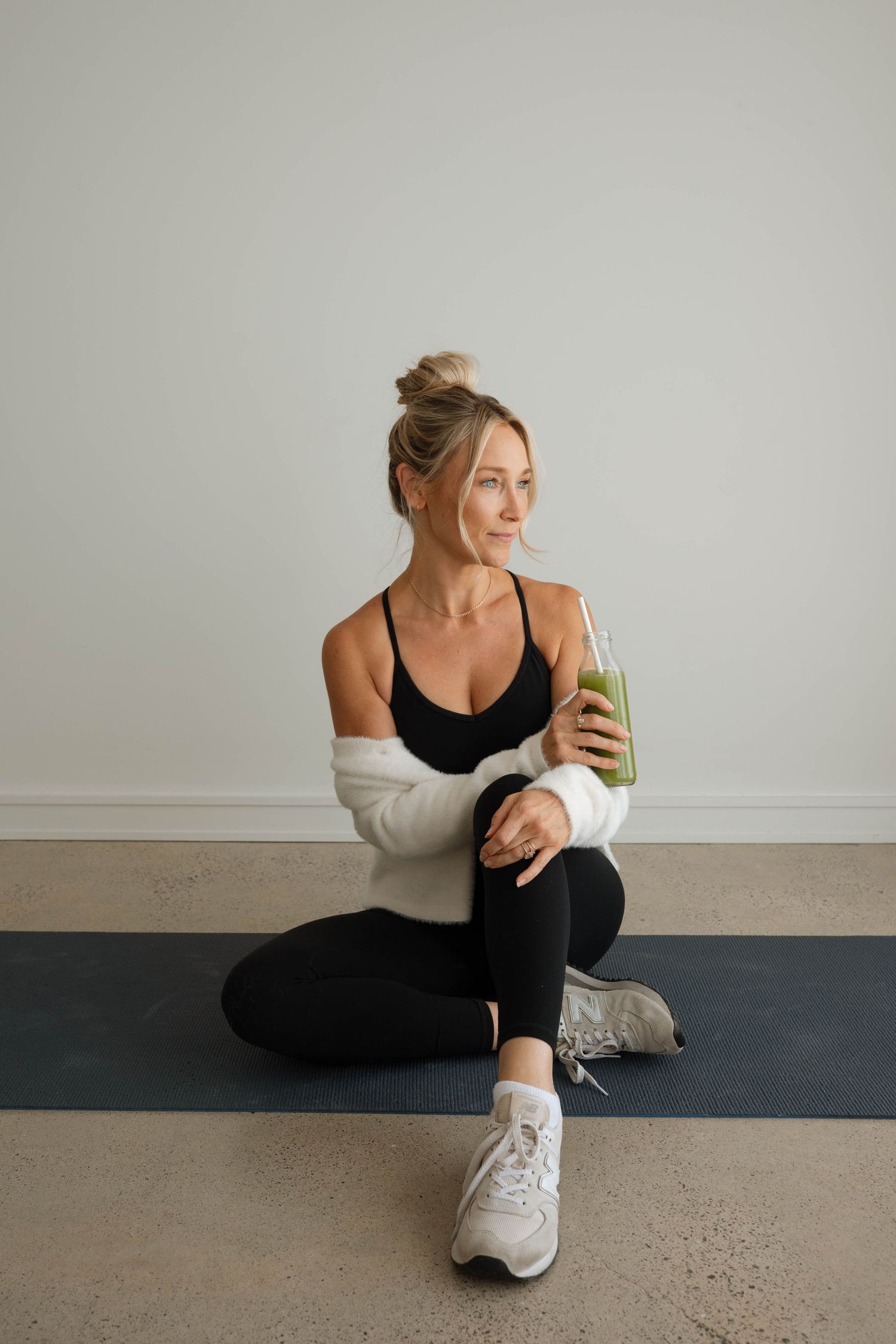 Fit blonde woman in black activewear sitting on yoga mat holding a green health drink.