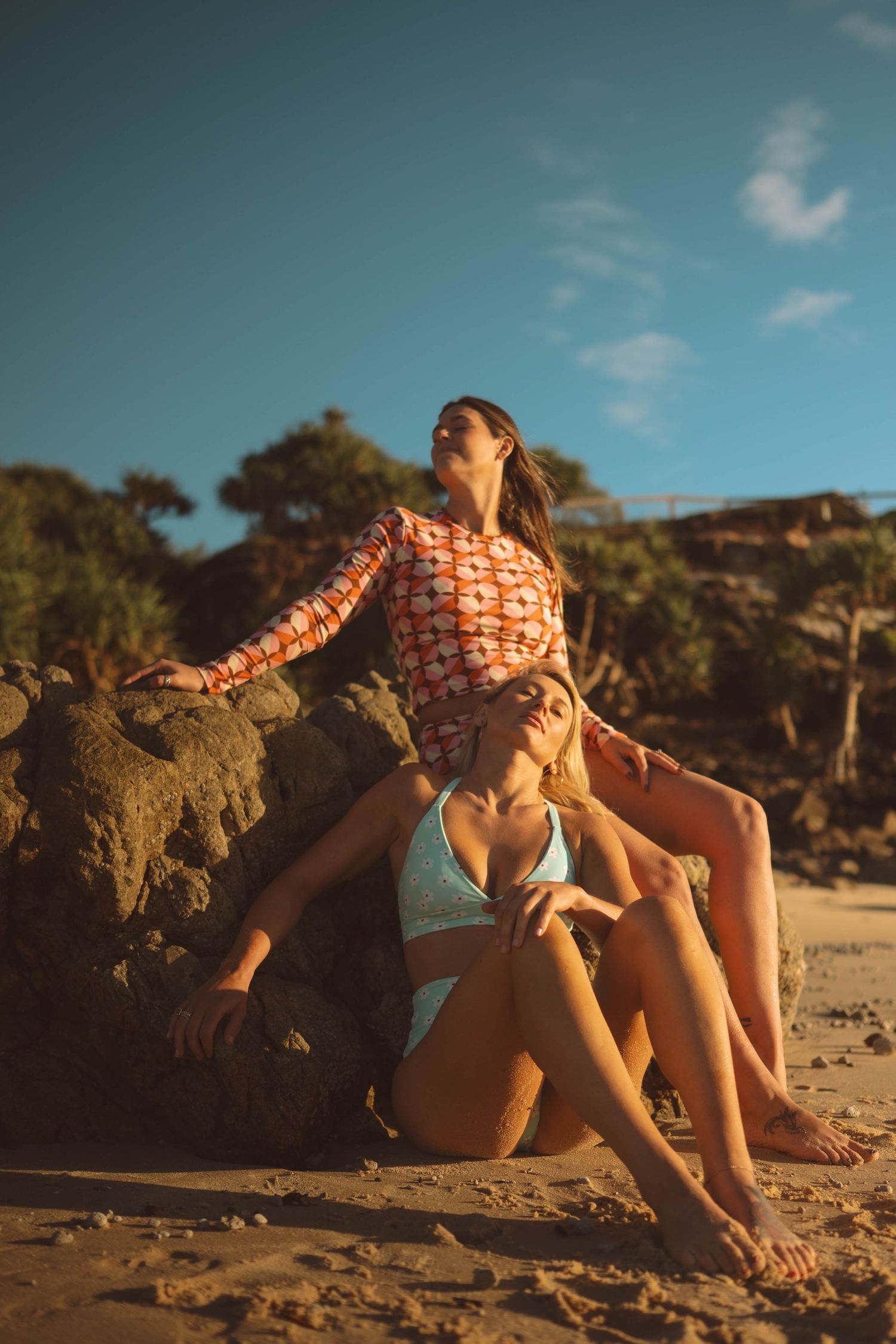 Two women in colorful swimwear posing against beach rocks under blue sky at sunset.