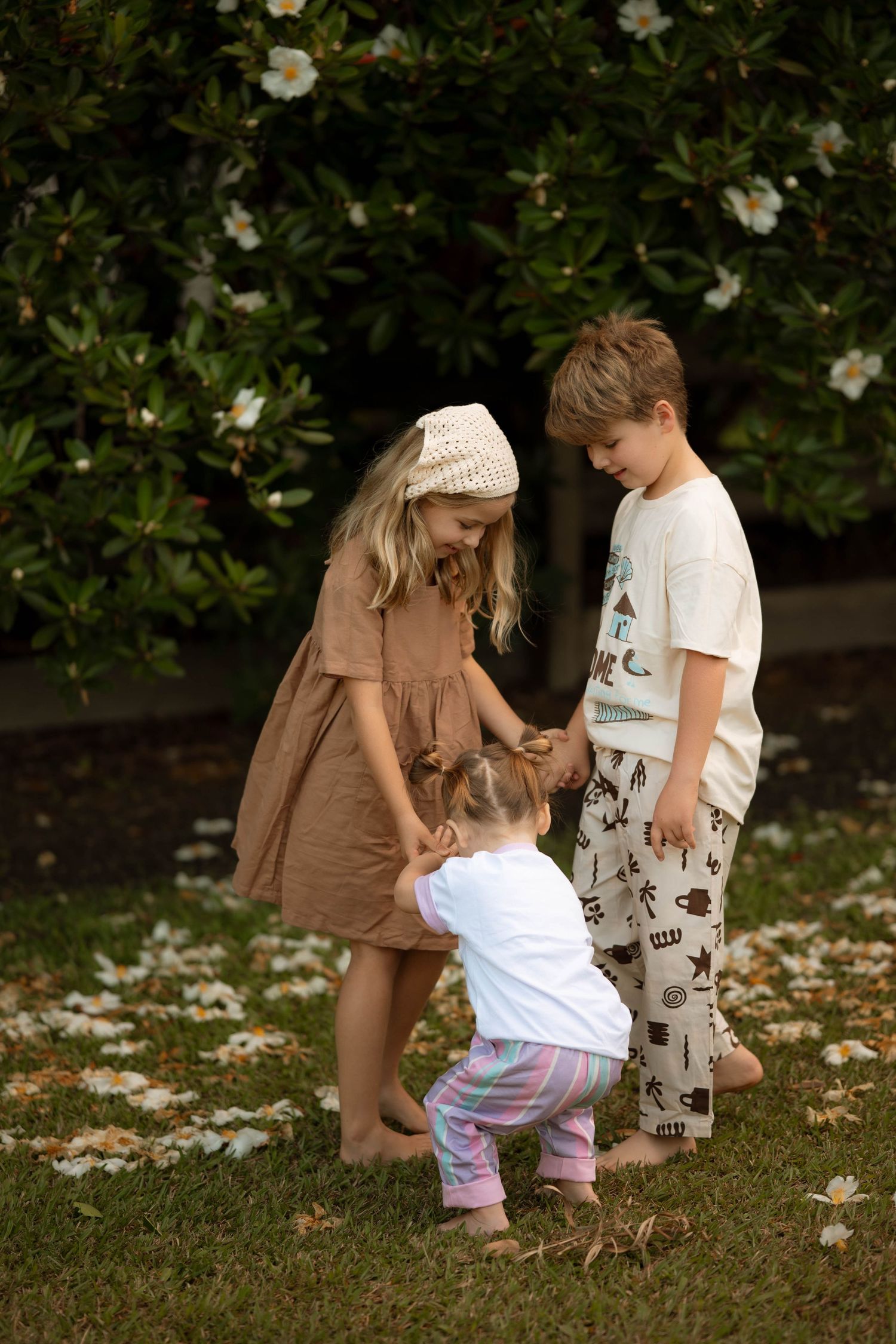 Three young children interact playfully on grass near white flowering bushes in natural light.