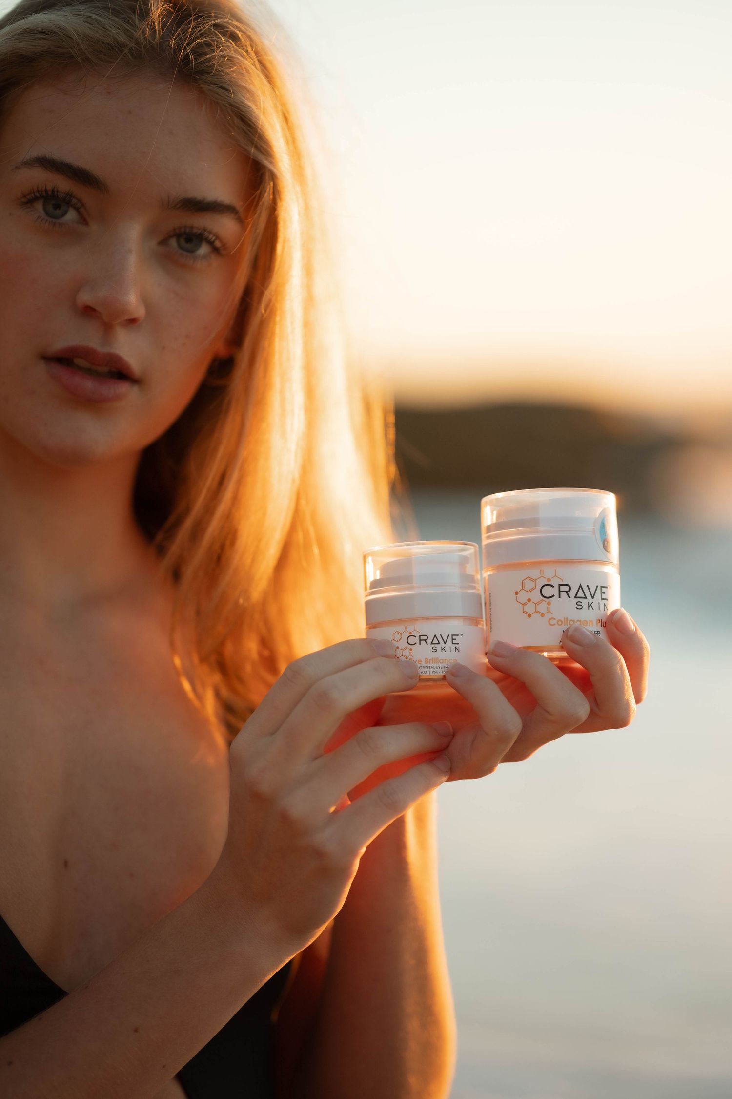 Blonde woman holding two Crave skincare products at sunset beach.