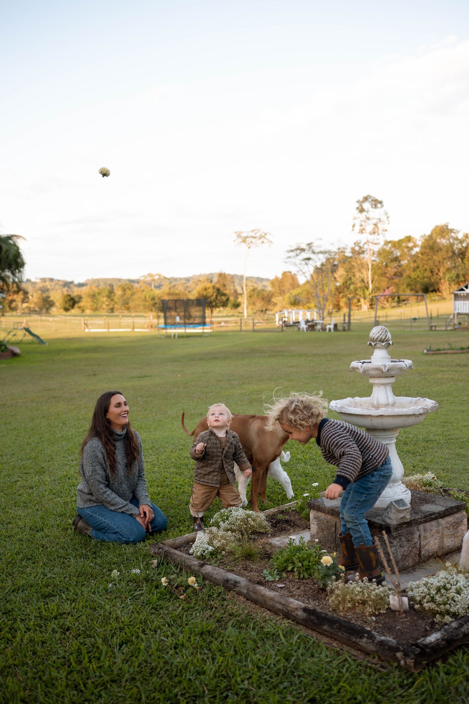 Woman sits on grass while two young children play near a stone fountain on a lush green farm property.