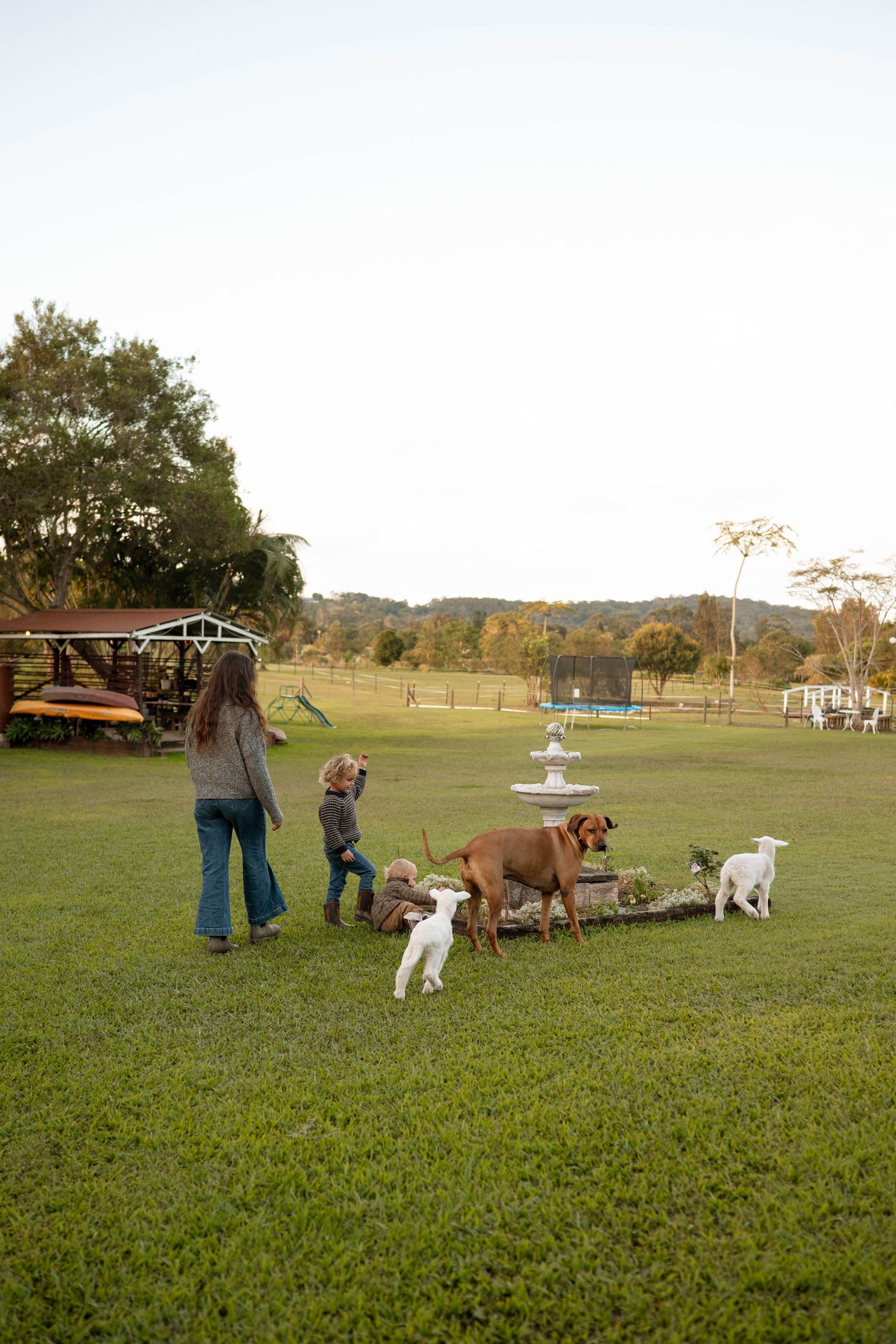 Woman and child playing with dogs and goats near a fountain in a lush green farm yard.
