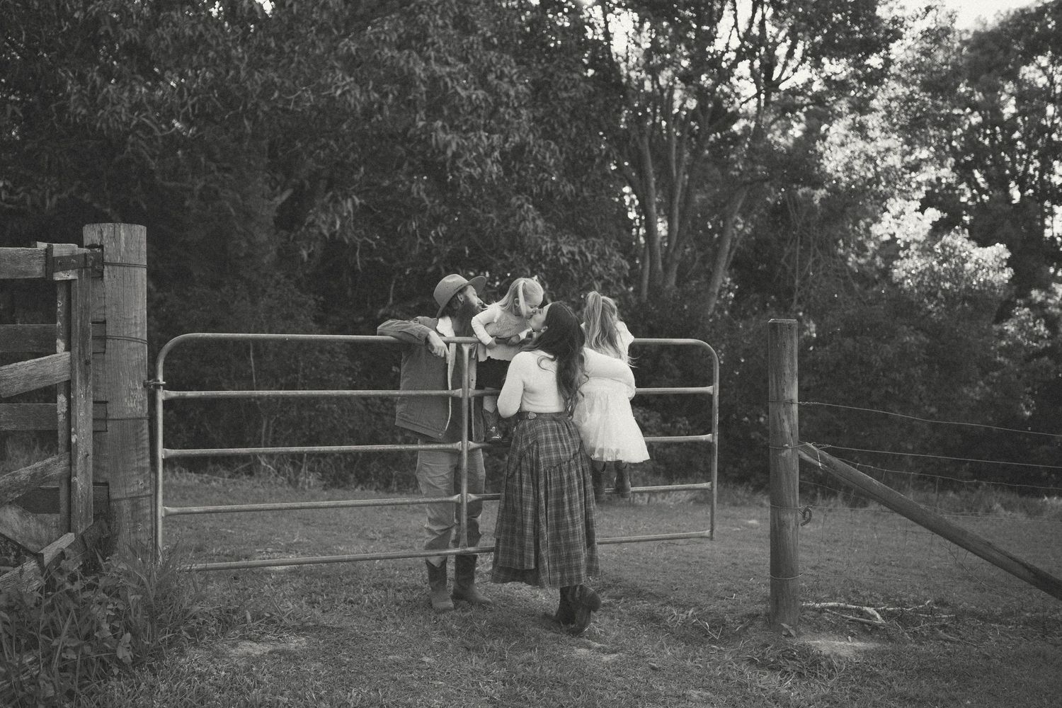 Black and white photo of two people in rural attire leaning on a farm gate surrounded by trees.
