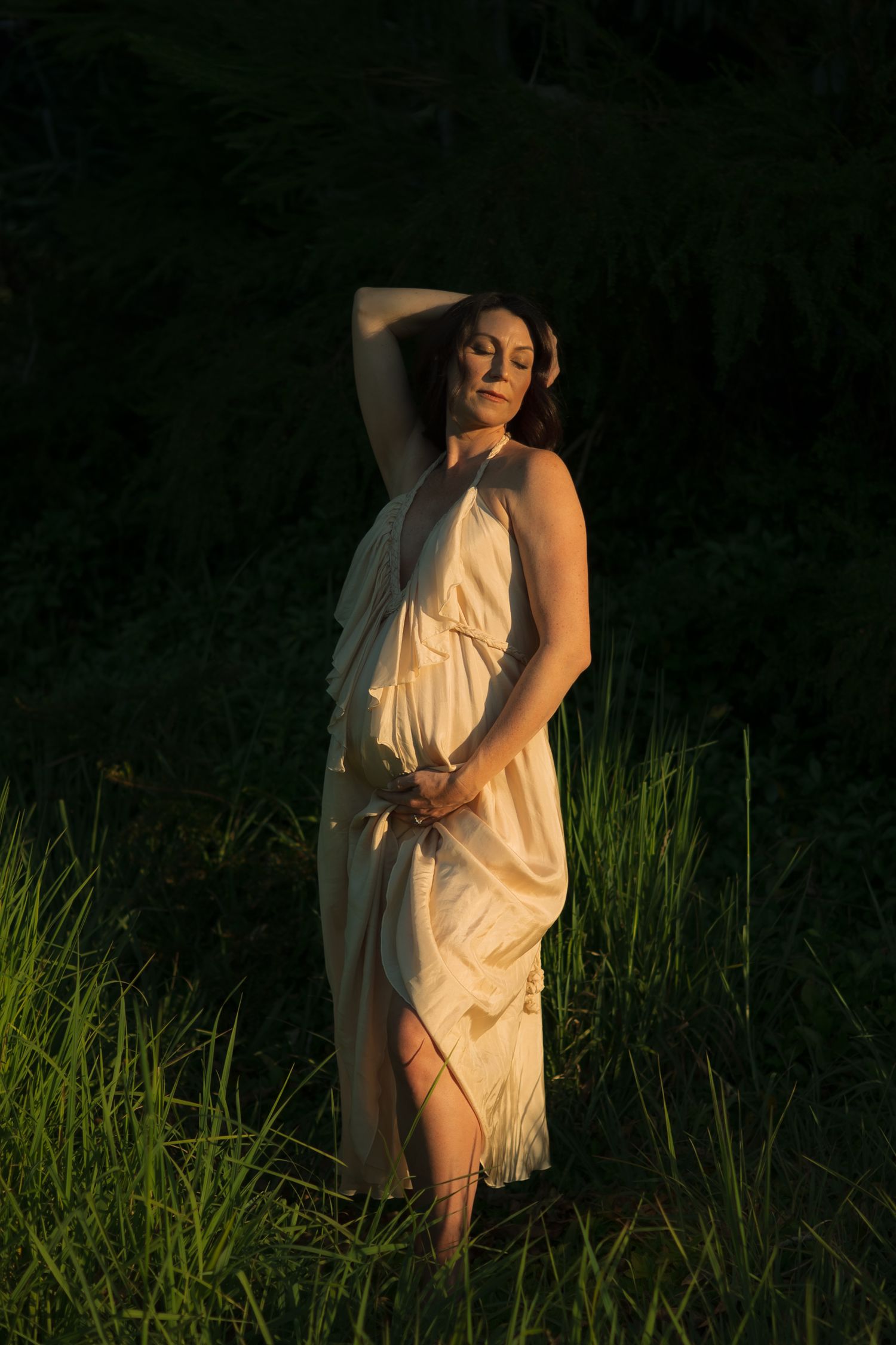 Pregnant woman in cream dress poses gracefully in golden hour light among lush green grass.