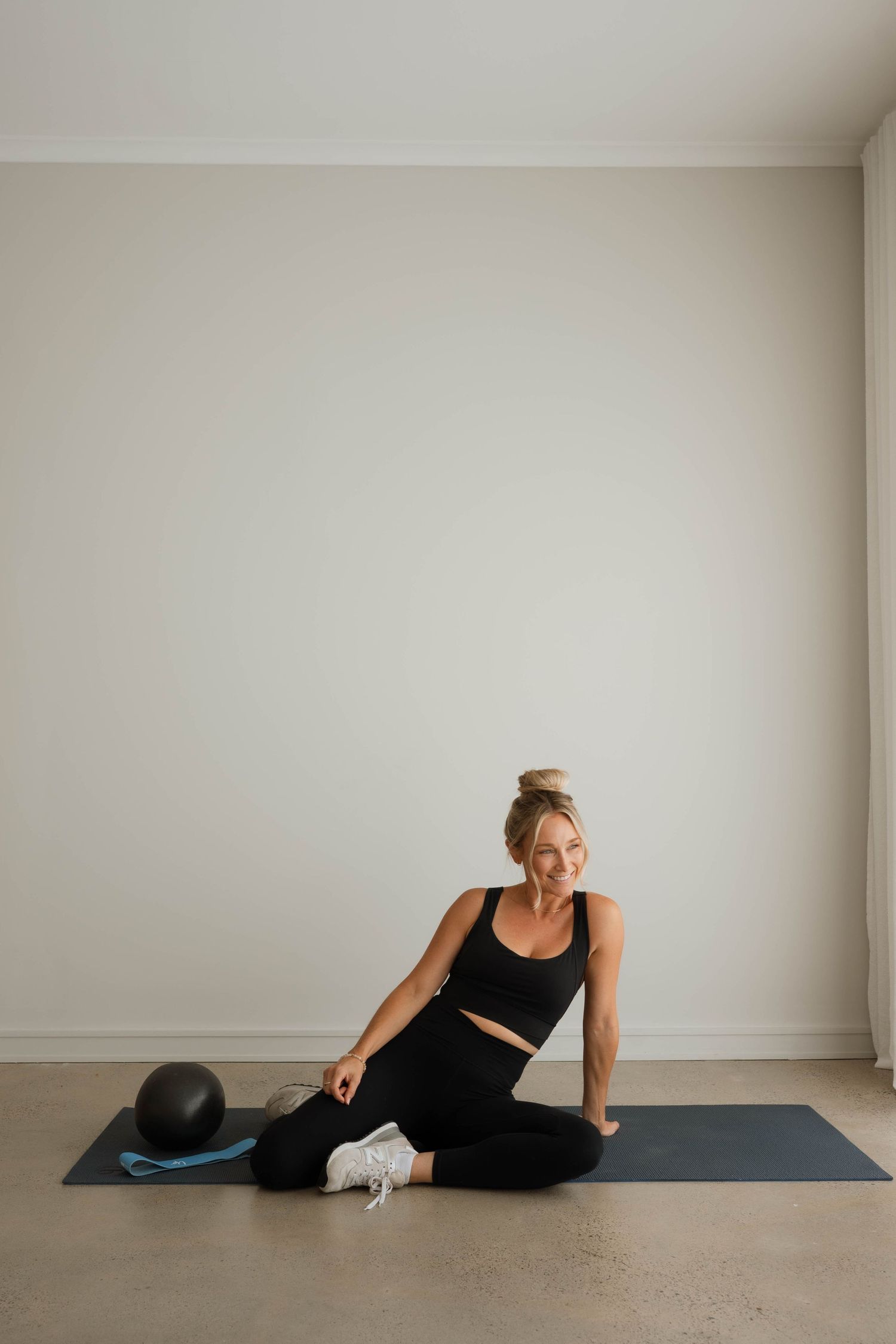 Fit woman in black activewear sitting on yoga mat with medicine ball in bright studio.