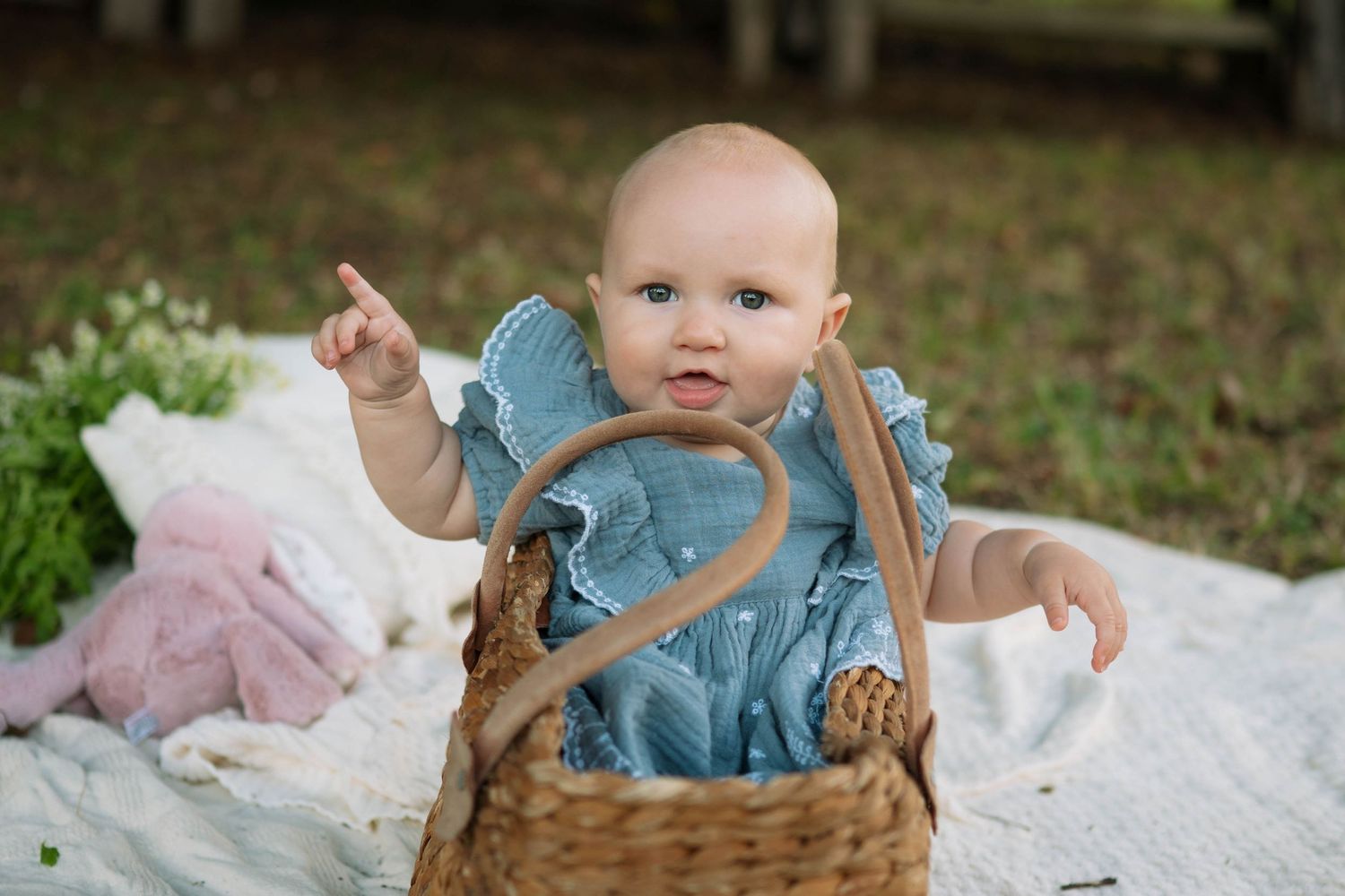 Cute baby in a blue ruffled dress sits in a wicker basket on a blanket, pointing one finger upward.