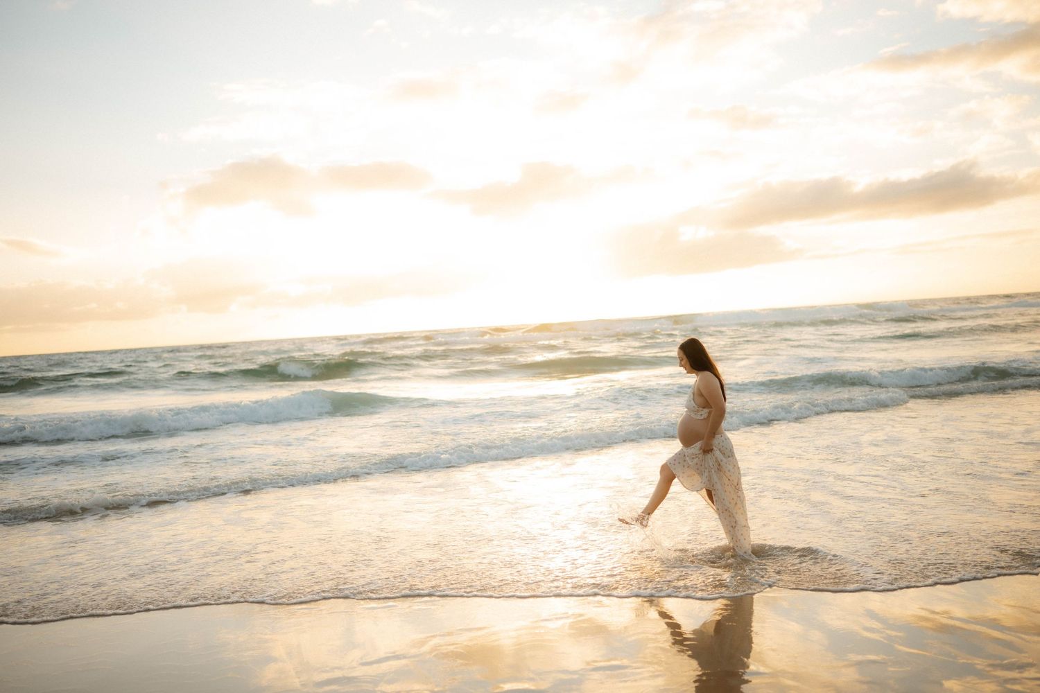 Expectant mother in flowing white dress strolling through shallow ocean waves during a warm beach sunset.