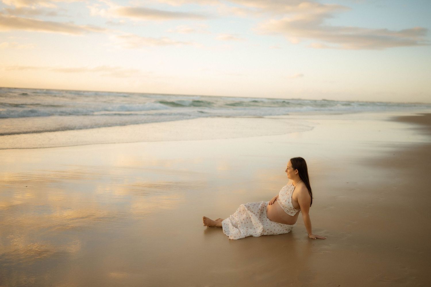 Serene pregnant woman sitting on reflective beach sand at sunset, looking peacefully toward ocean waves.