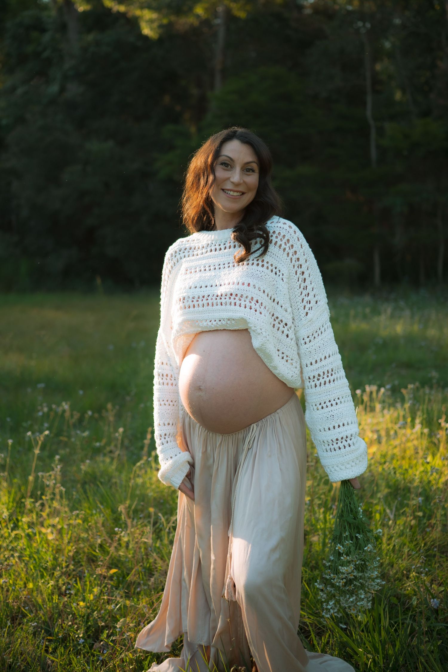 Smiling pregnant woman in white knit sweater holding her bare bump in a sunny green meadow.