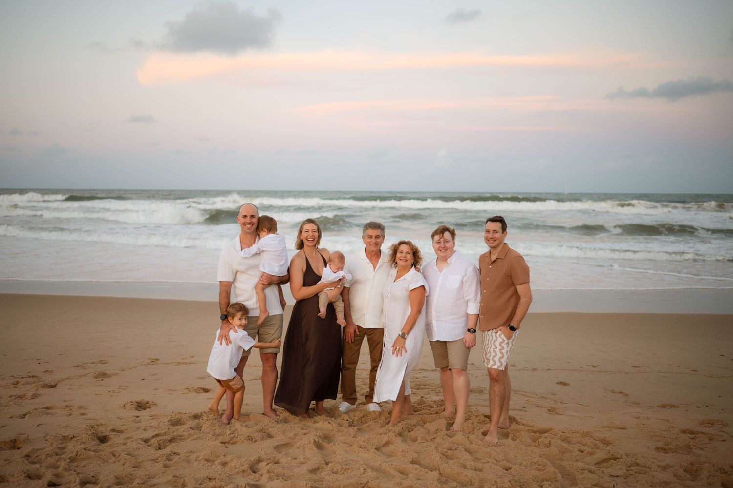 Extended family of seven posing together on a beach with ocean waves and pastel sunset sky behind them.