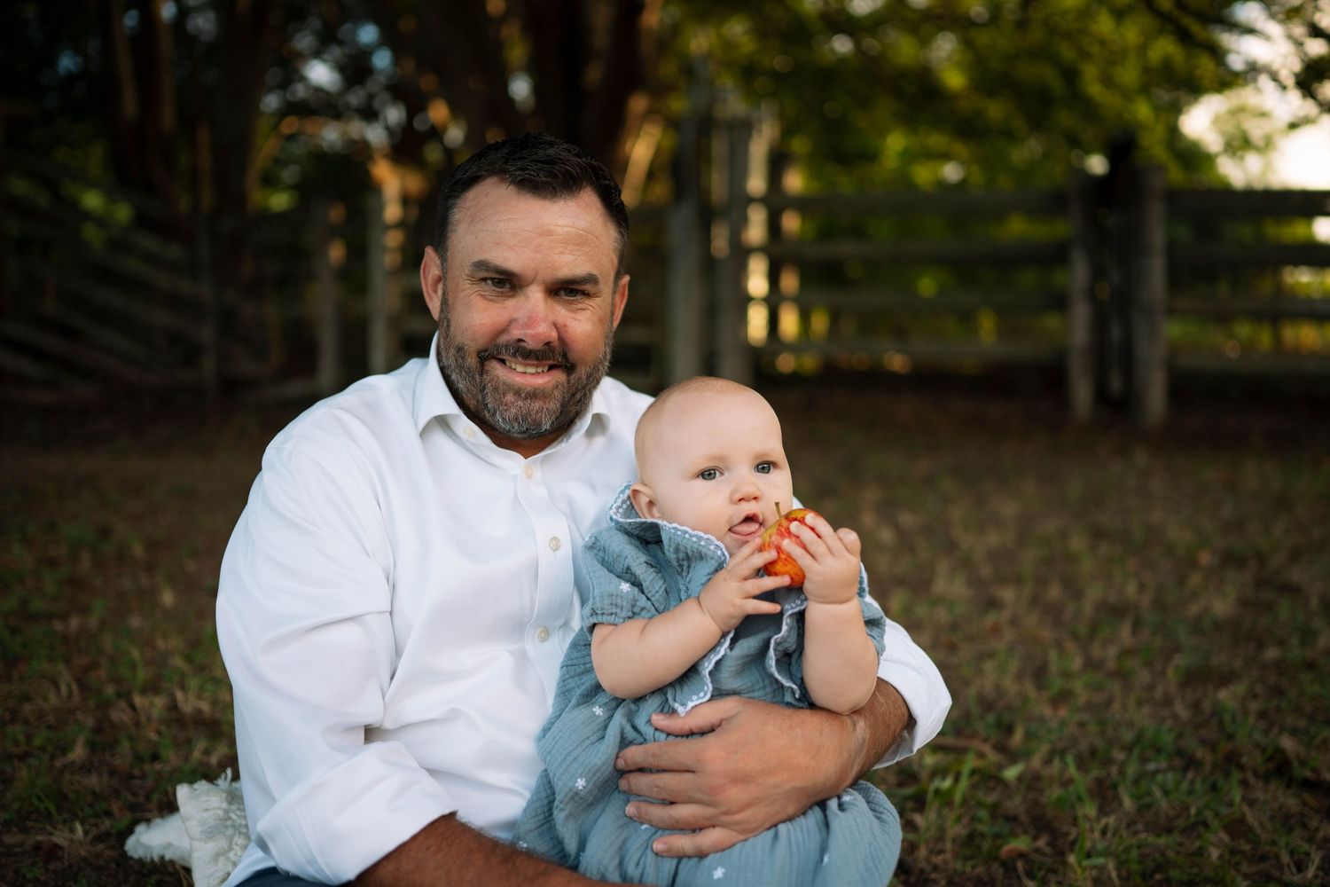 Smiling bearded father in a white shirt sits outdoors holding his baby in a blue outfit chewing a toy.