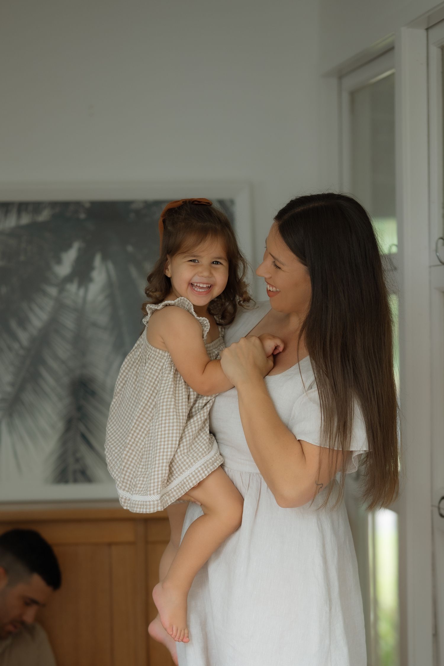 Smiling mother lifting happy toddler daughter in bright home with palm tree artwork.