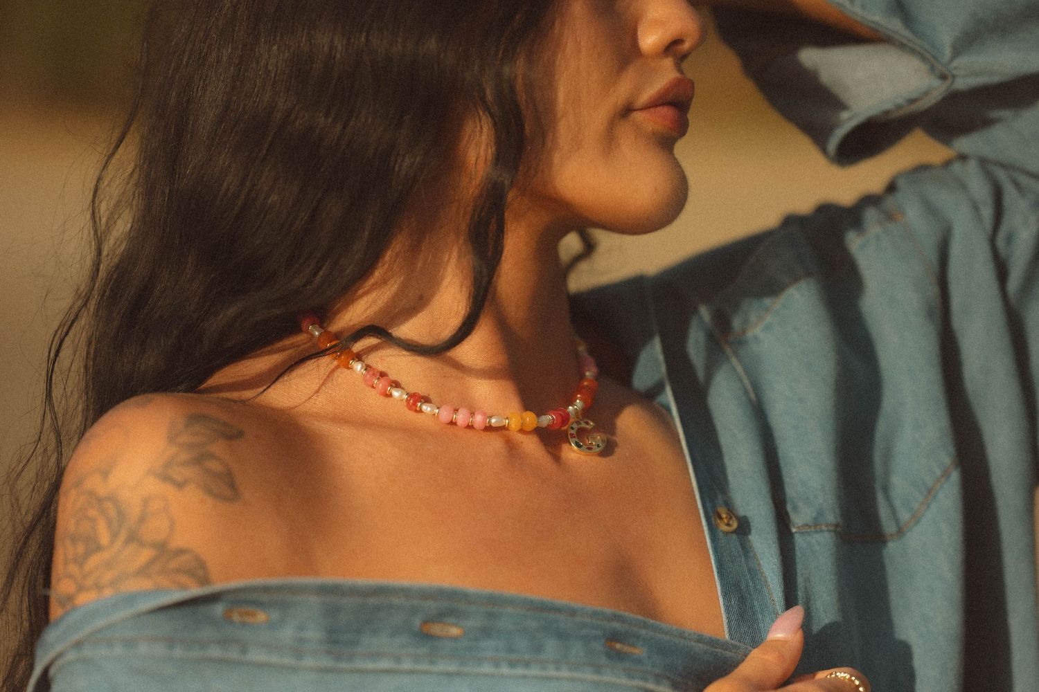 Woman wearing colorful beaded necklace and denim top with floral tattoo on shoulder.