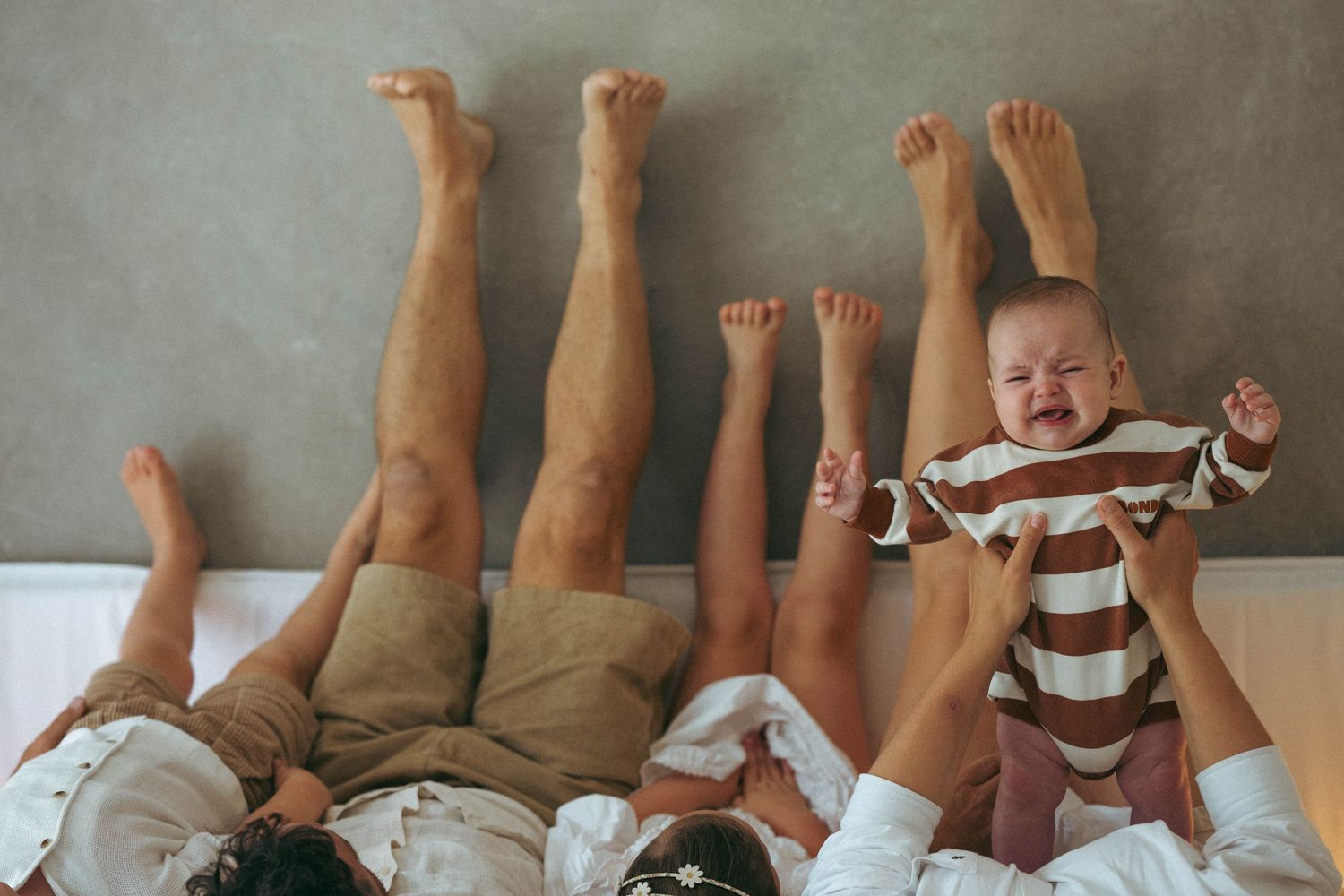 Family lying on bed with legs raised, parents lifting happy baby in striped onesie, fun playful moment.