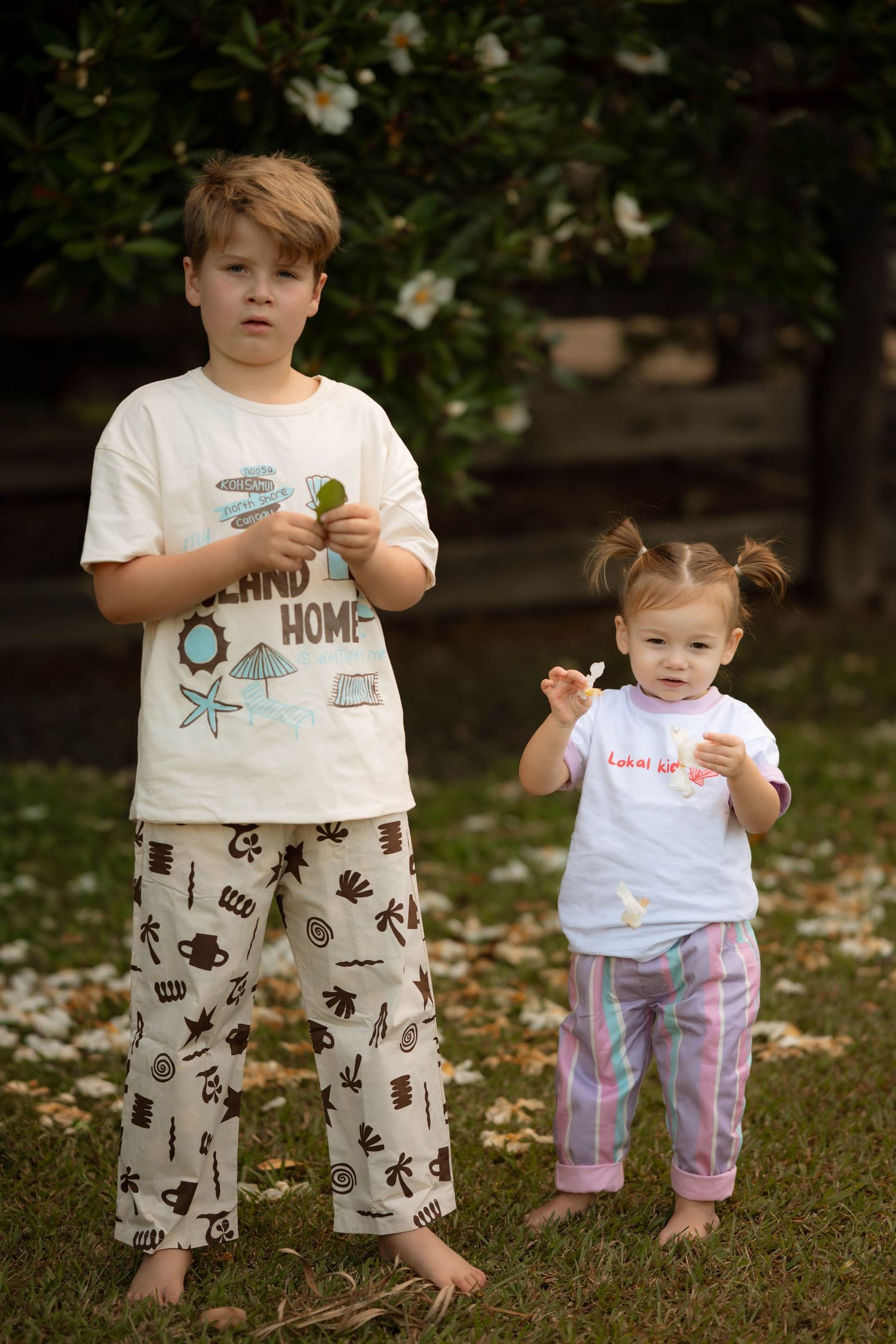 Two children in pajamas standing on grass, older boy holding an object, toddler girl with pigtails beside him.