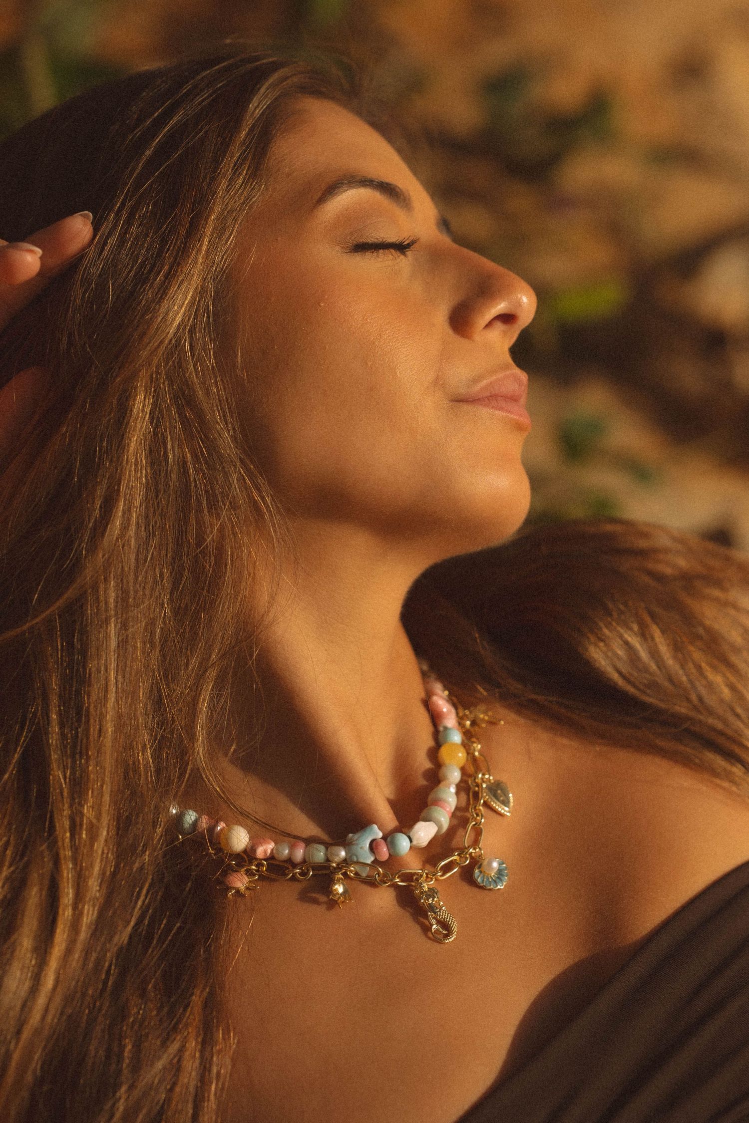 Woman with eyes closed wearing colorful beaded charm necklaces, bathed in warm golden sunlight.