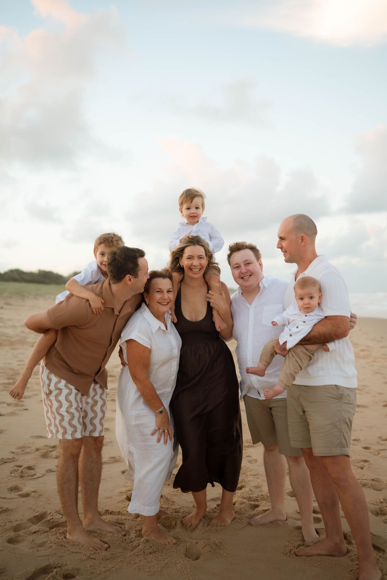 Large multigenerational family of seven smiling together on a sandy beach with soft cloudy sky backdrop.