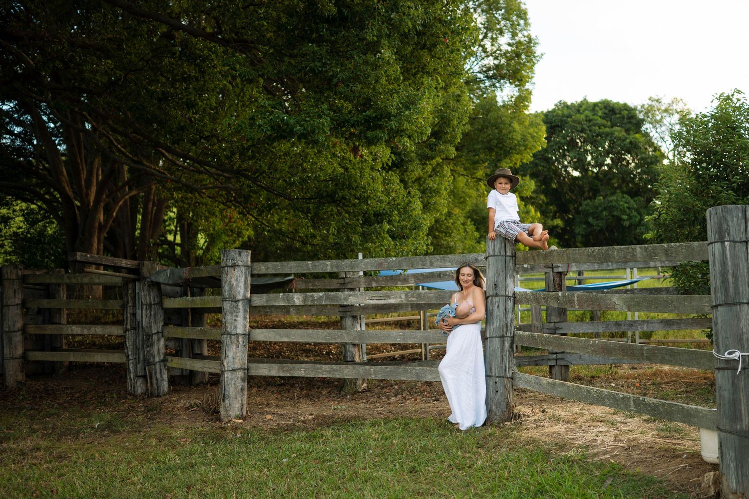 A bride in a white dress stands by a rustic wooden fence while a boy sits on top in a lush green farm setting.