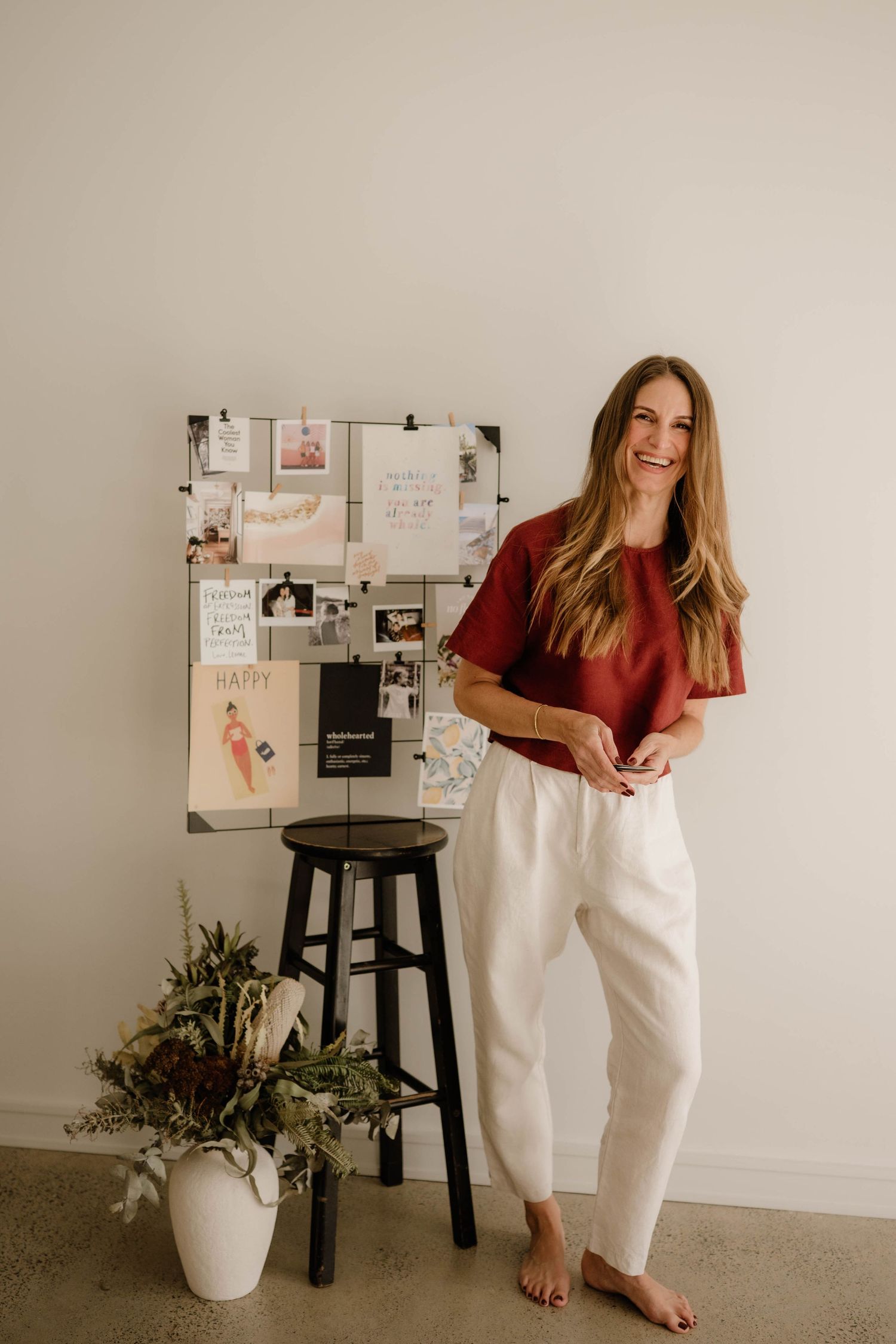 Smiling woman in red top and white pants standing beside mood board with dried flowers.