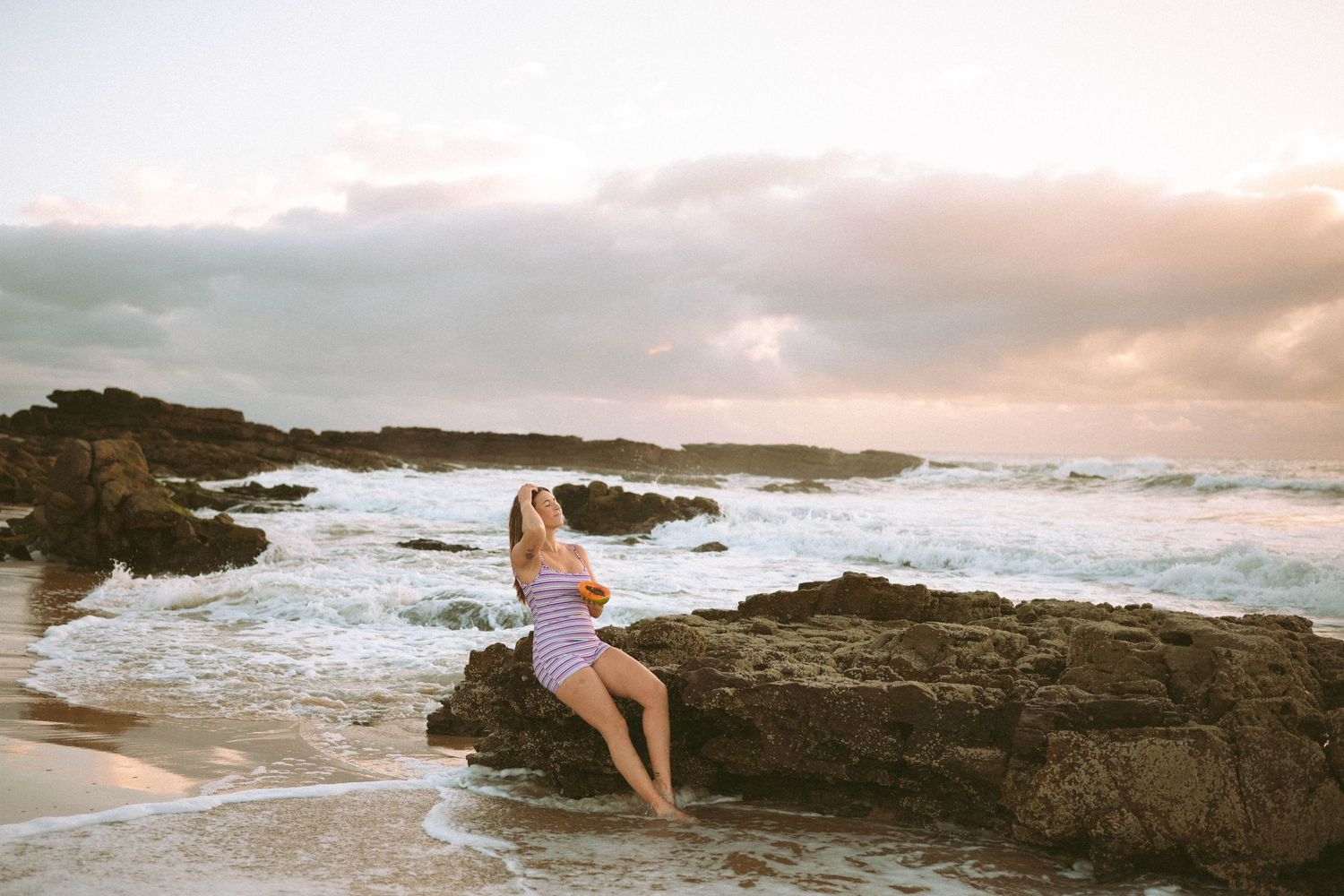 Woman in striped dress sitting on coastal rocks at sunset with ocean waves behind her.