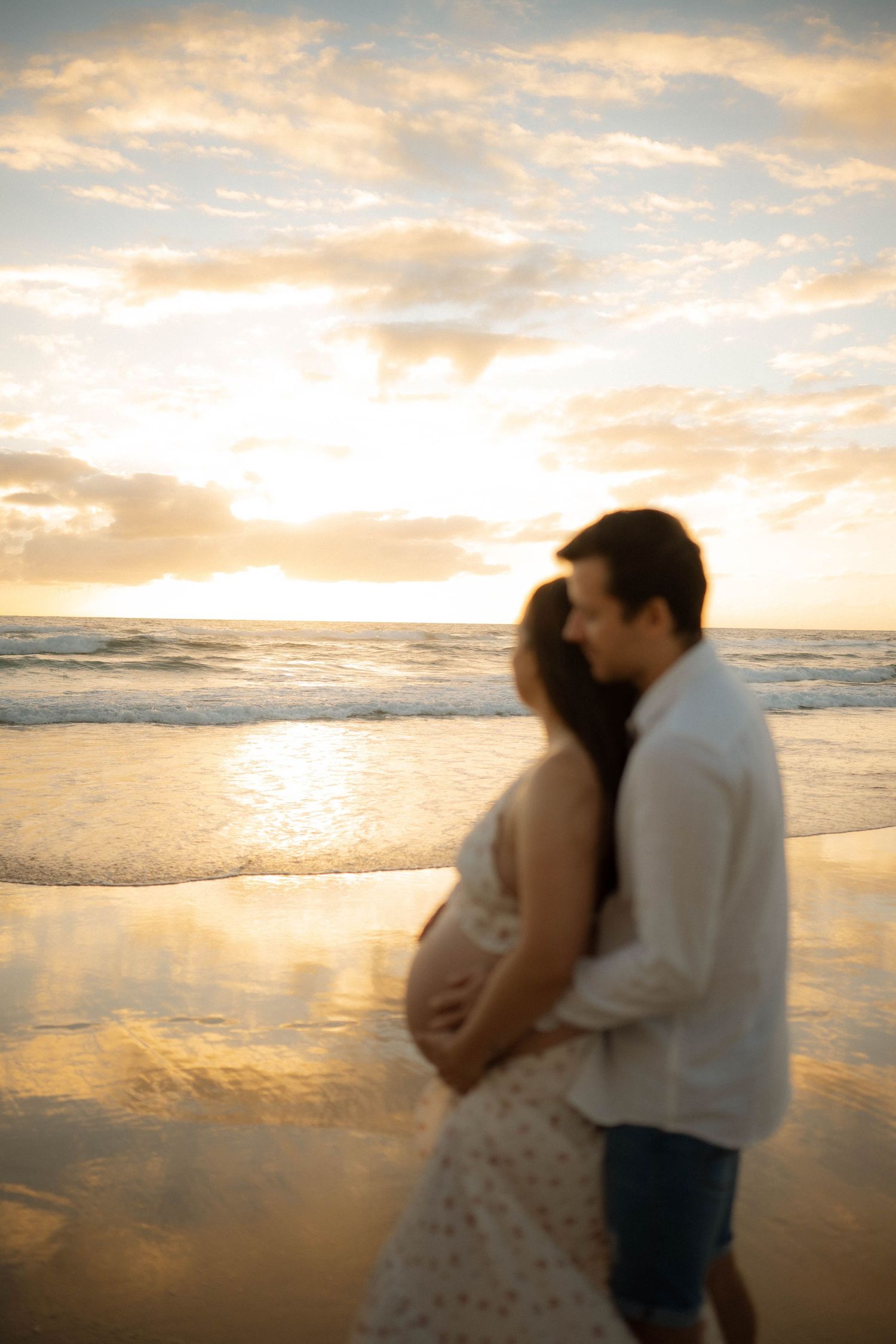 Silhouetted pregnant couple embracing at beach sunset, man holding baby bump in warm golden light.