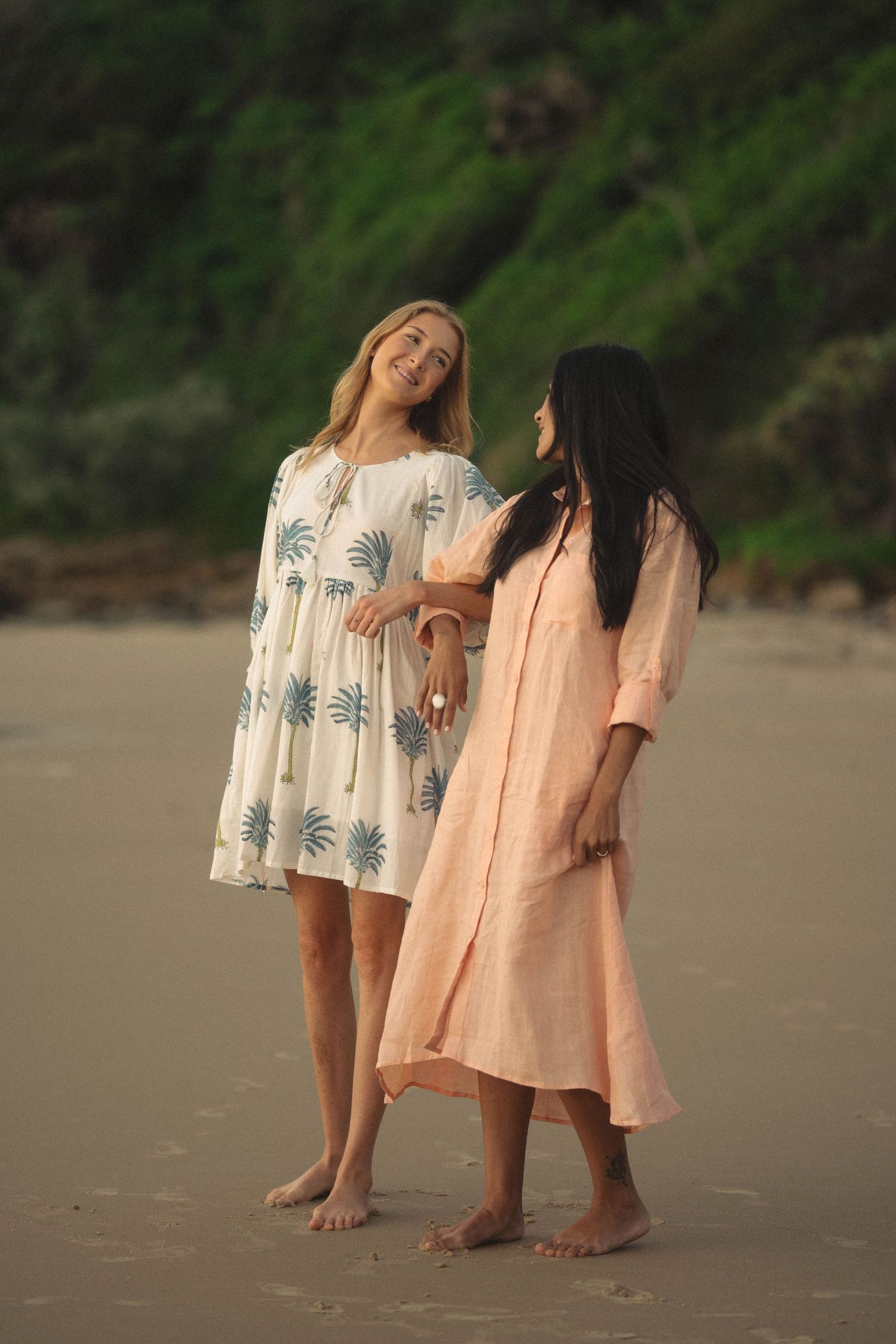Two women in flowy dresses stand barefoot on a sandy beach, smiling and posing together near green cliffs.