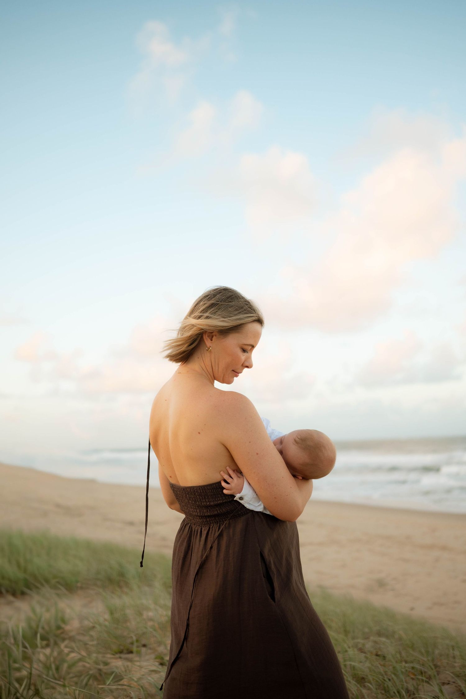 Mother in brown strapless dress breastfeeding newborn on beach at sunset.