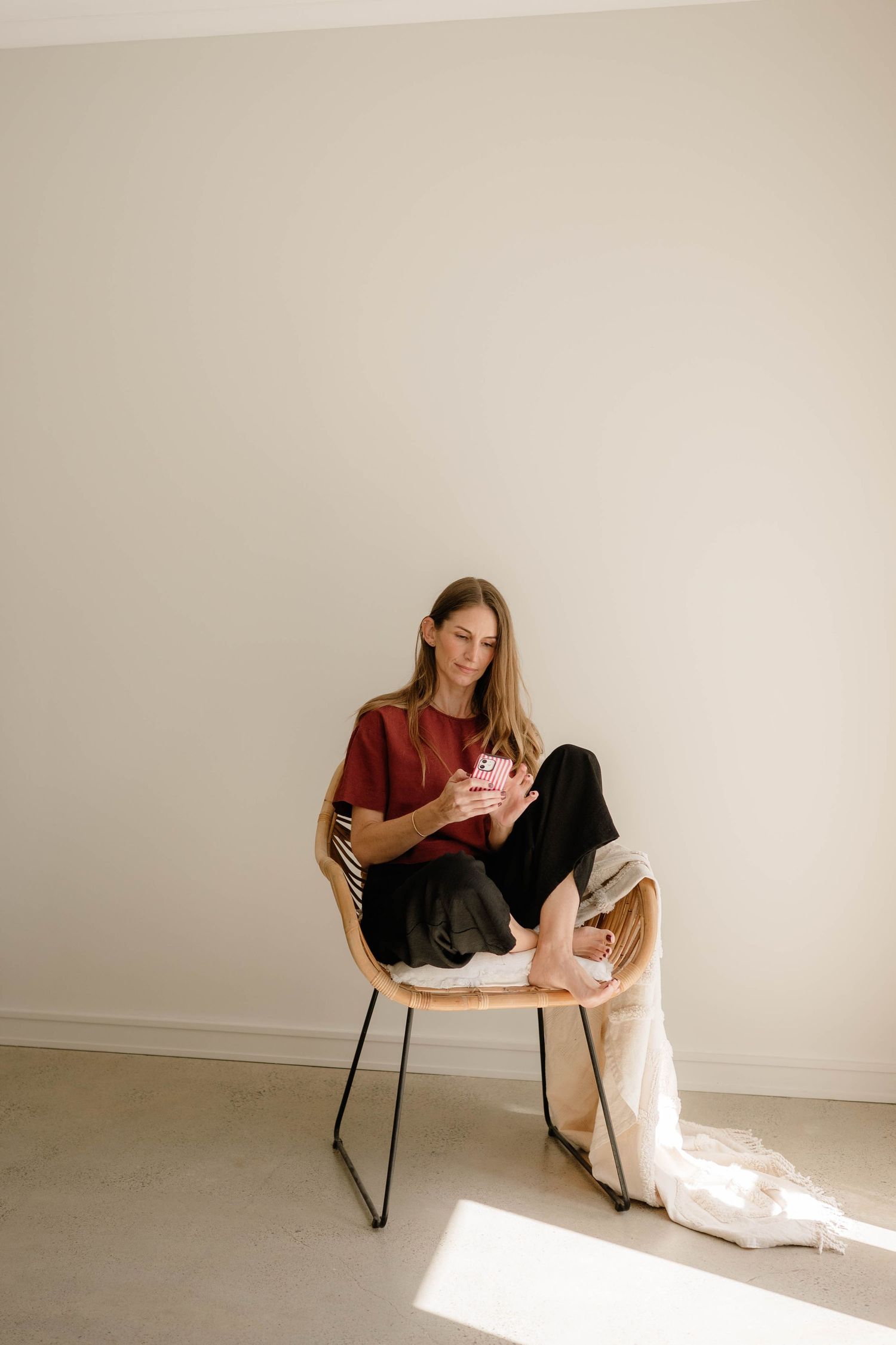 Young woman in red top and black pants sitting in rattan chair using pink smartphone.