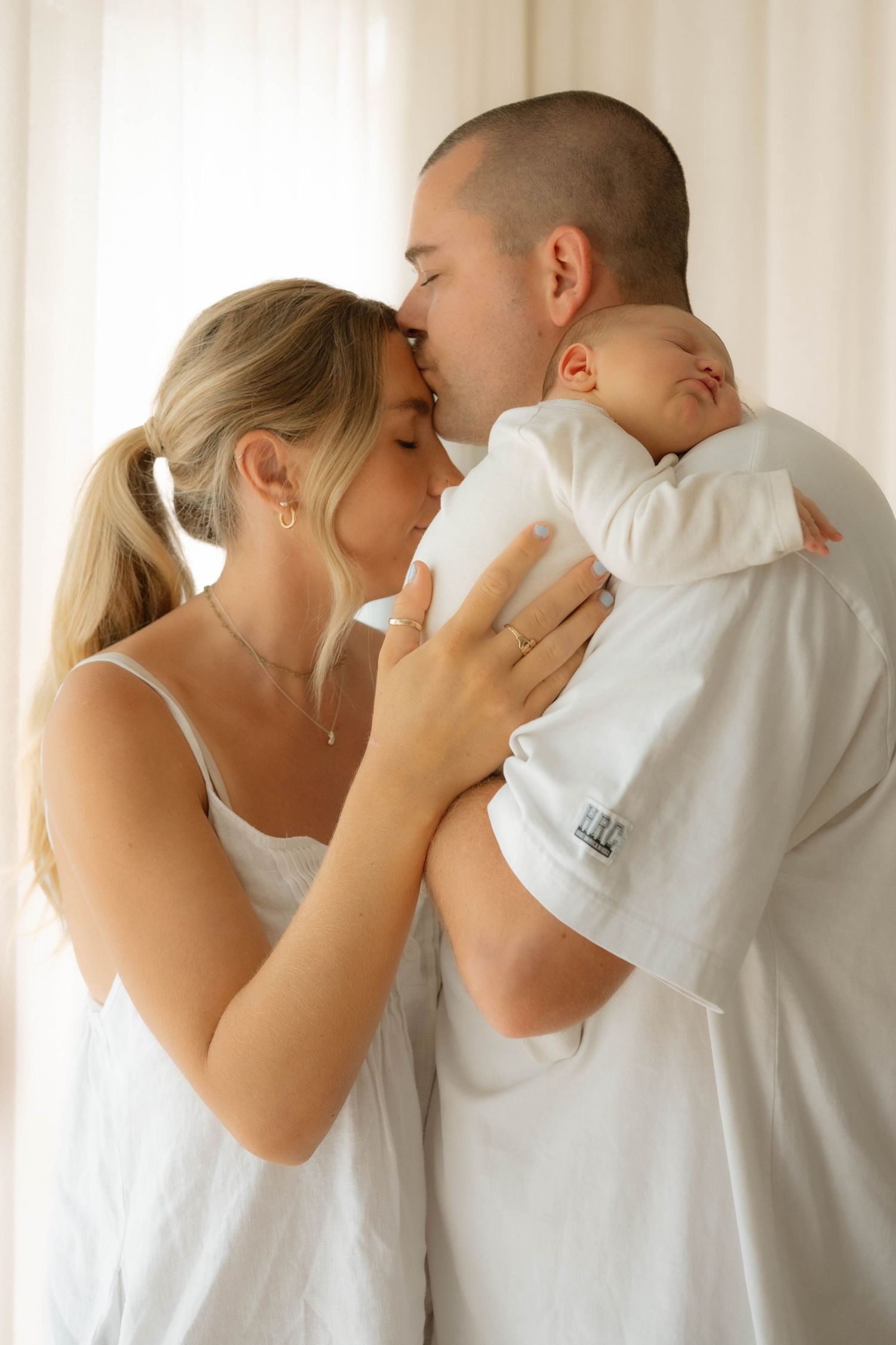 Parents kissing over newborn baby dressed in white against soft white curtain background.