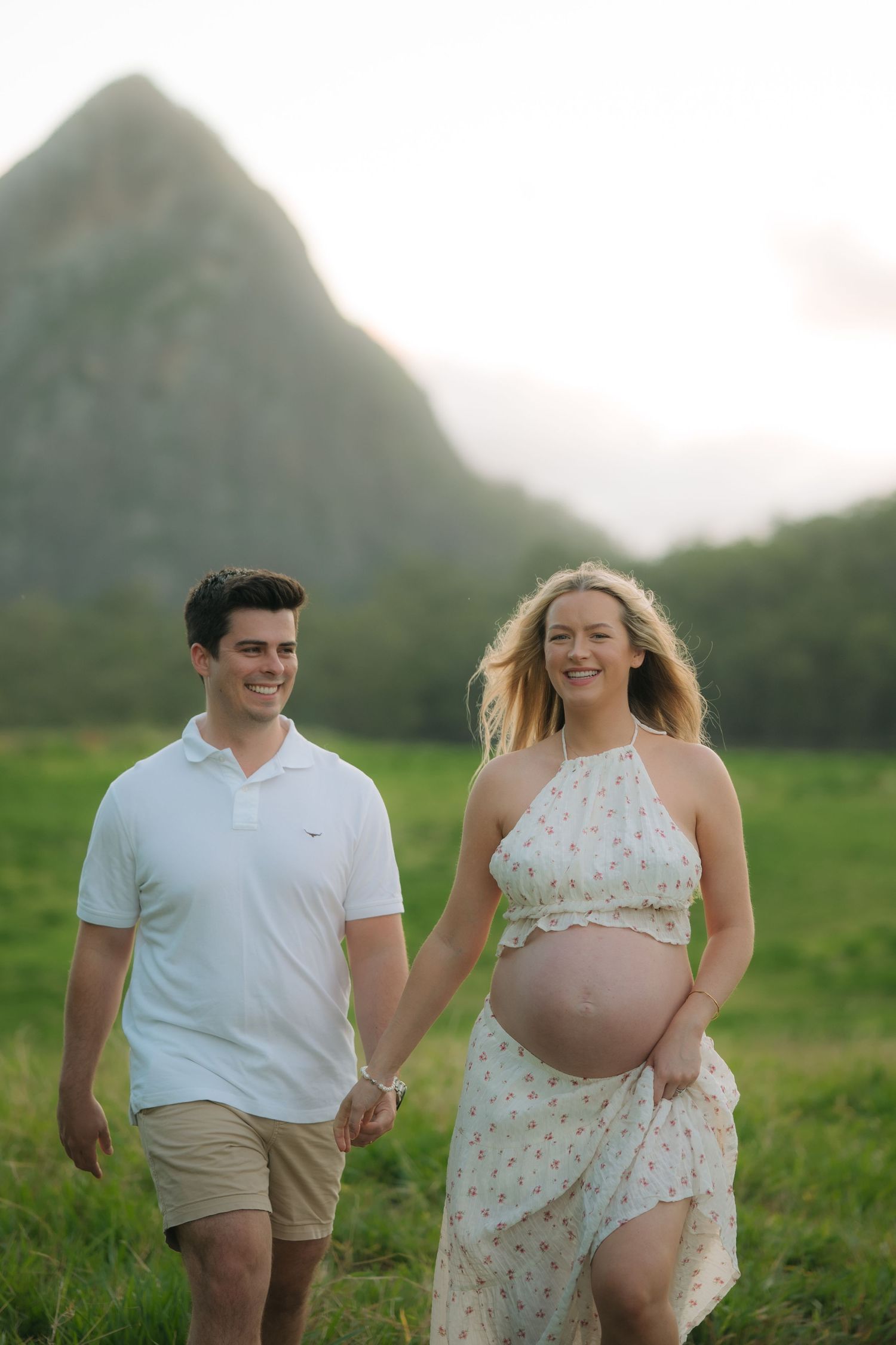 Smiling pregnant couple walks hand in hand through green field with mountain backdrop during golden hour.